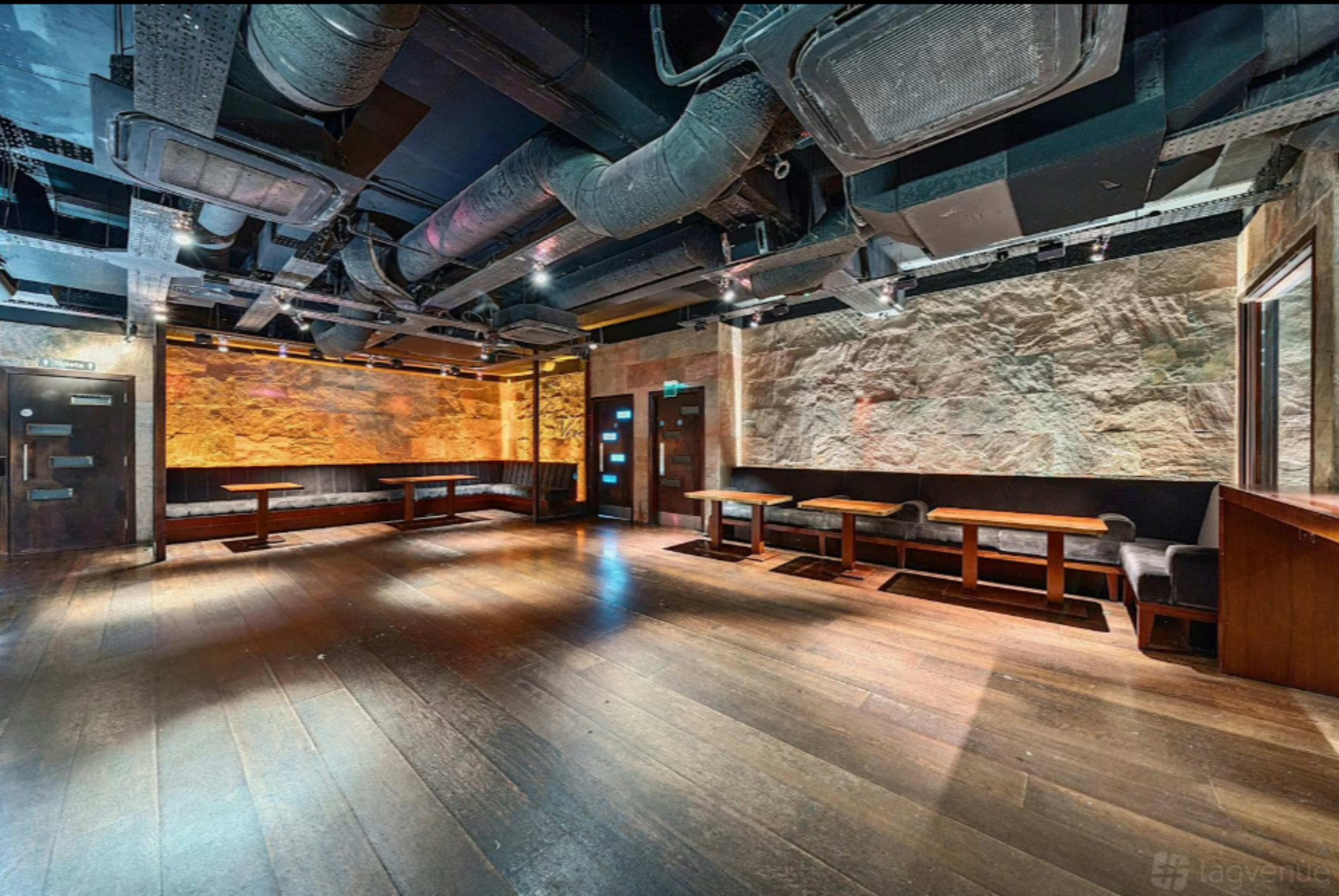 The image shows a spacious interior of a dimly lit venue featuring wooden flooring, exposed ductwork, and several tables arranged in a modern layout.