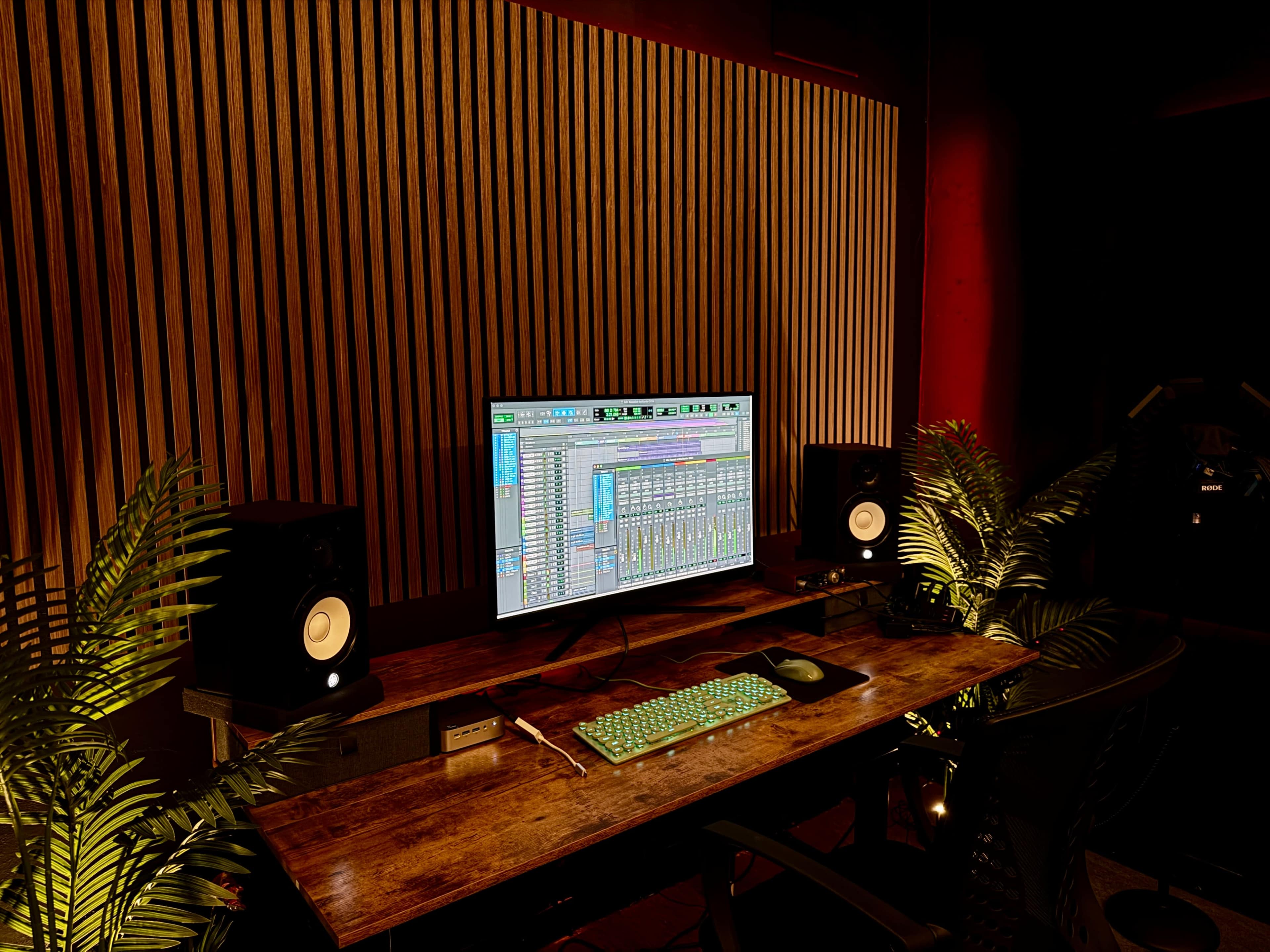 A music production workspace features a computer monitor displaying sound mixing software, flanked by studio monitors and surrounded by greenery.