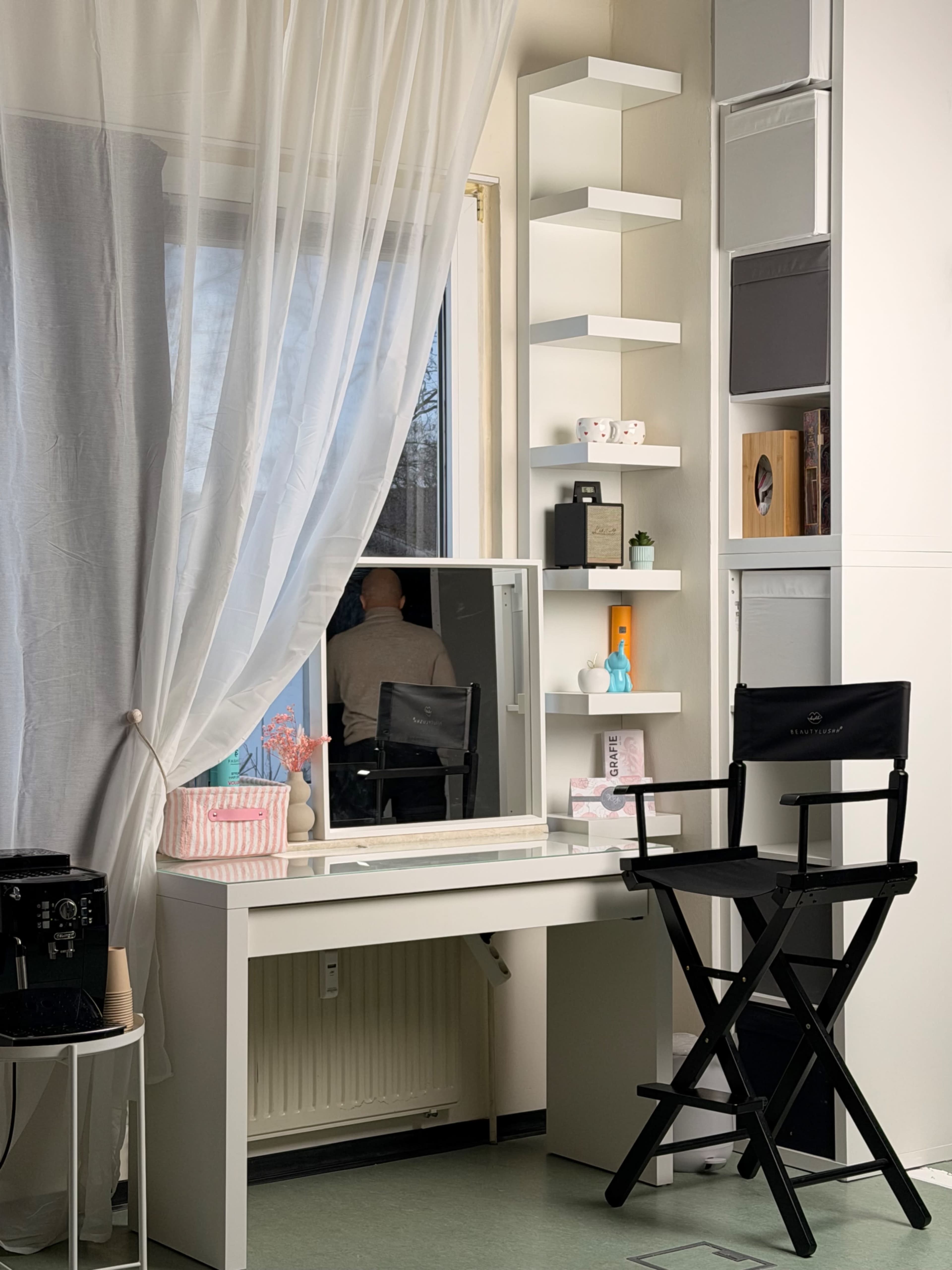 A bright room features a vanity with a mirror, a director's chair, and shelves displaying various decorative items, with sheer curtains framing a window.