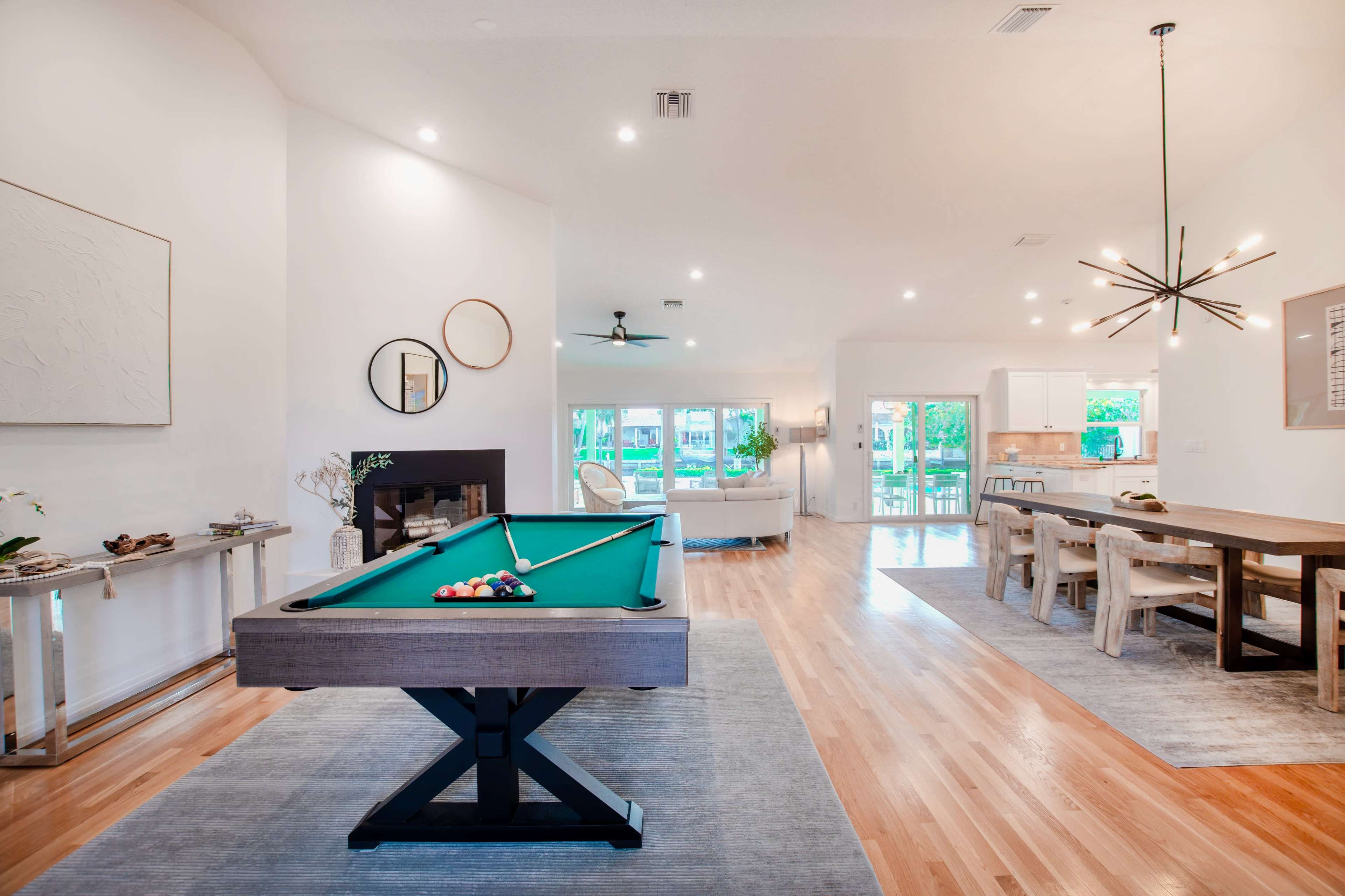 The image depicts a spacious, modern living area featuring a pool table, dining table, and large windows that allow natural light to fill the room.