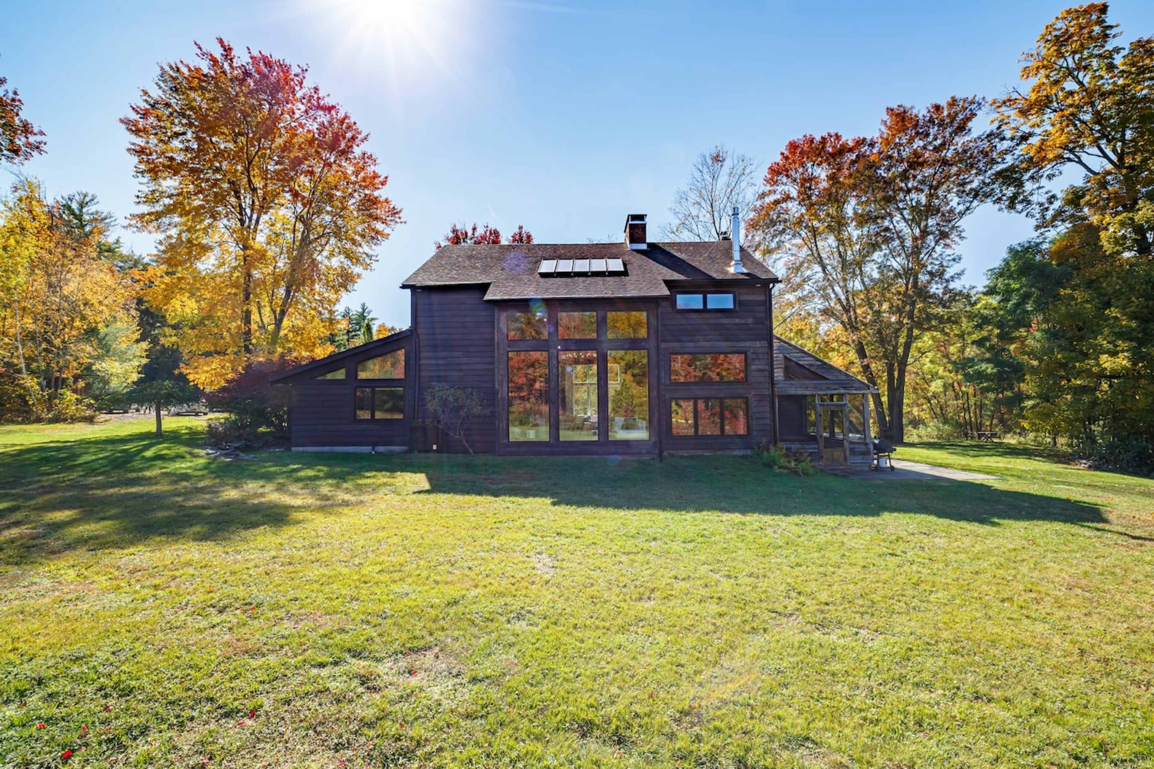 A large wooden house with large windows is surrounded by autumn foliage and a grassy area under clear blue skies.