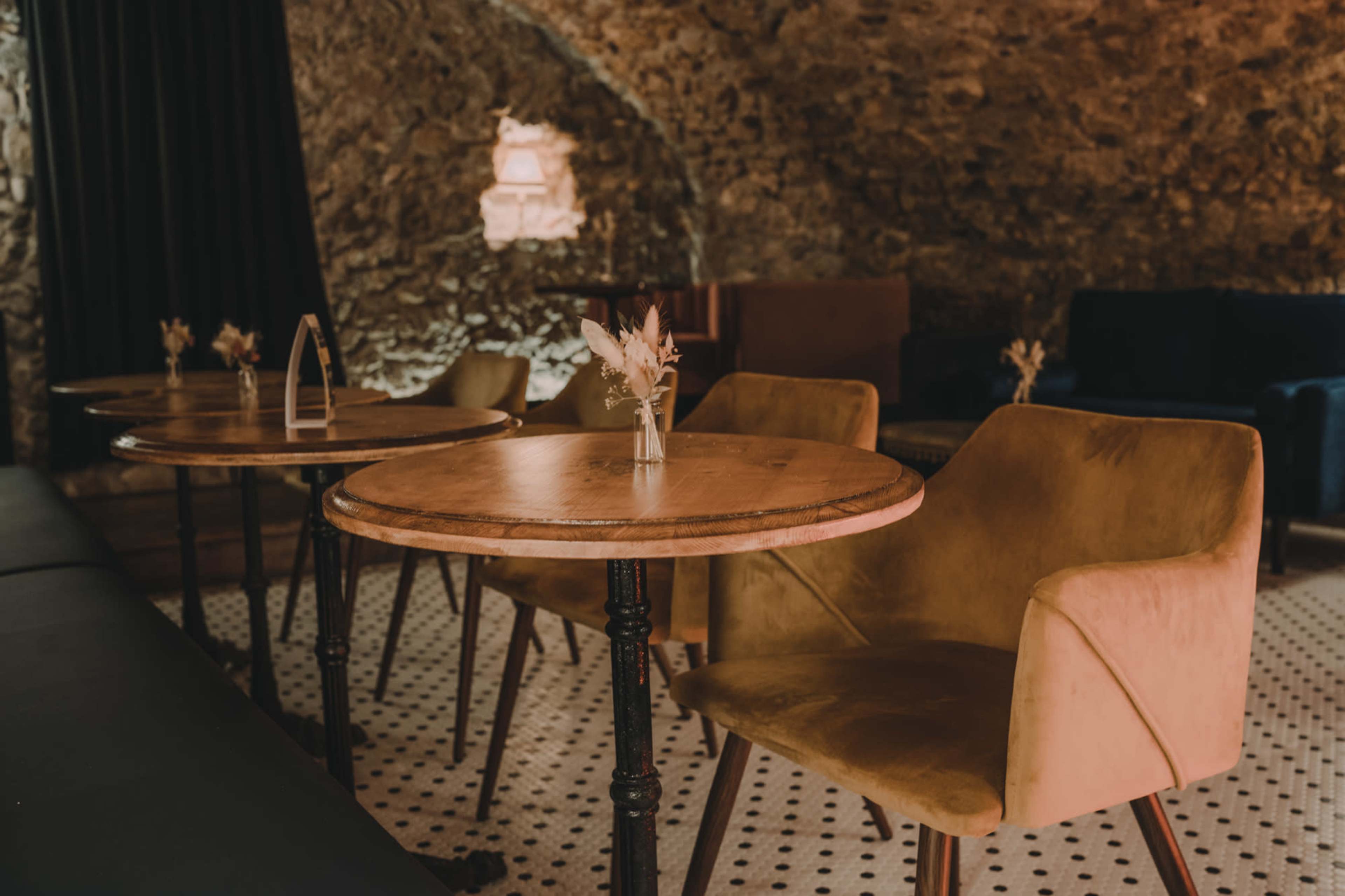 The image shows a cozy, underground lounge with stone walls, wooden tables, and upholstered chairs.