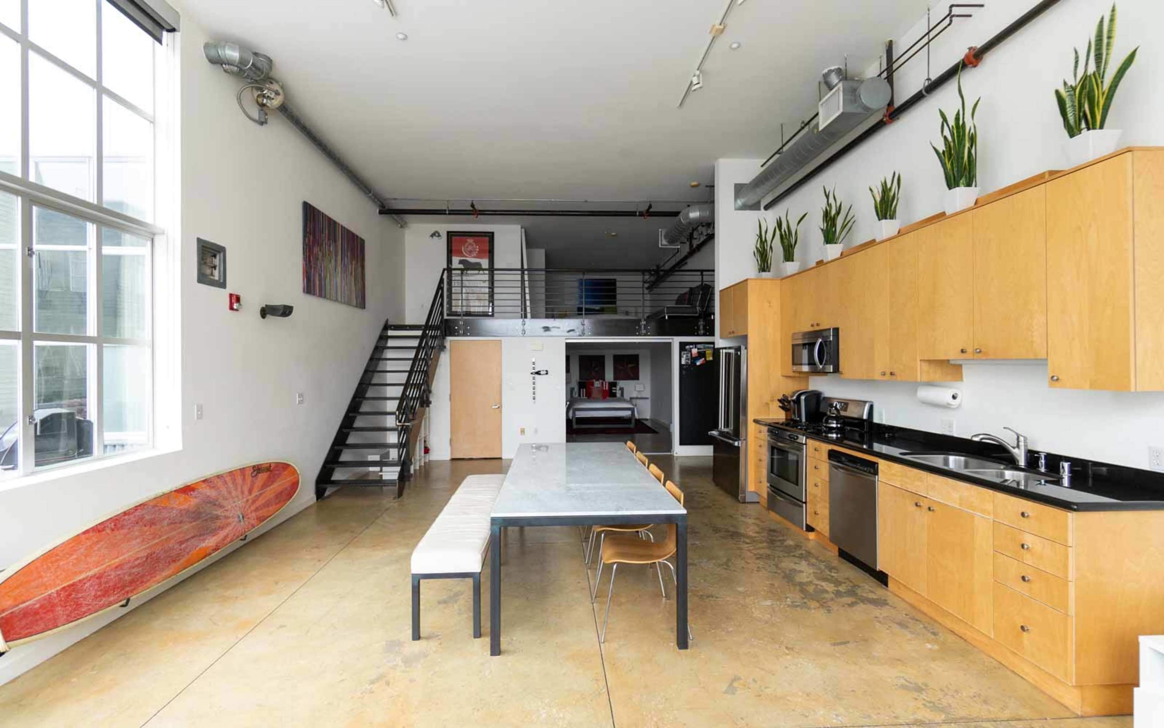 An open-concept kitchen and dining area featuring a long table, wooden cabinets, kitchen appliances, and a surfboard leaning against the wall.