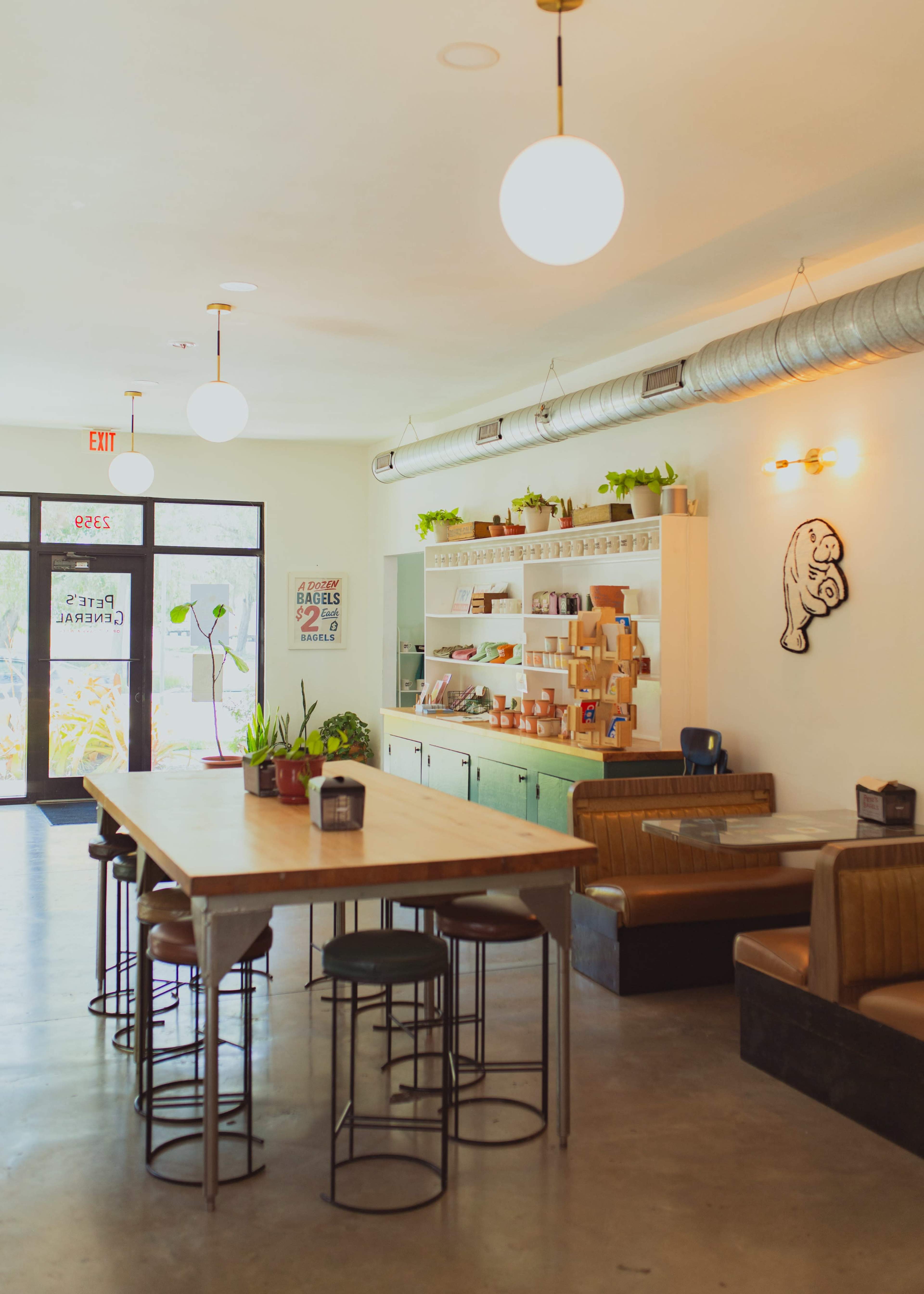 The image shows a modern café with a large communal table, seating areas, and a well-organized shelving unit displaying various items.