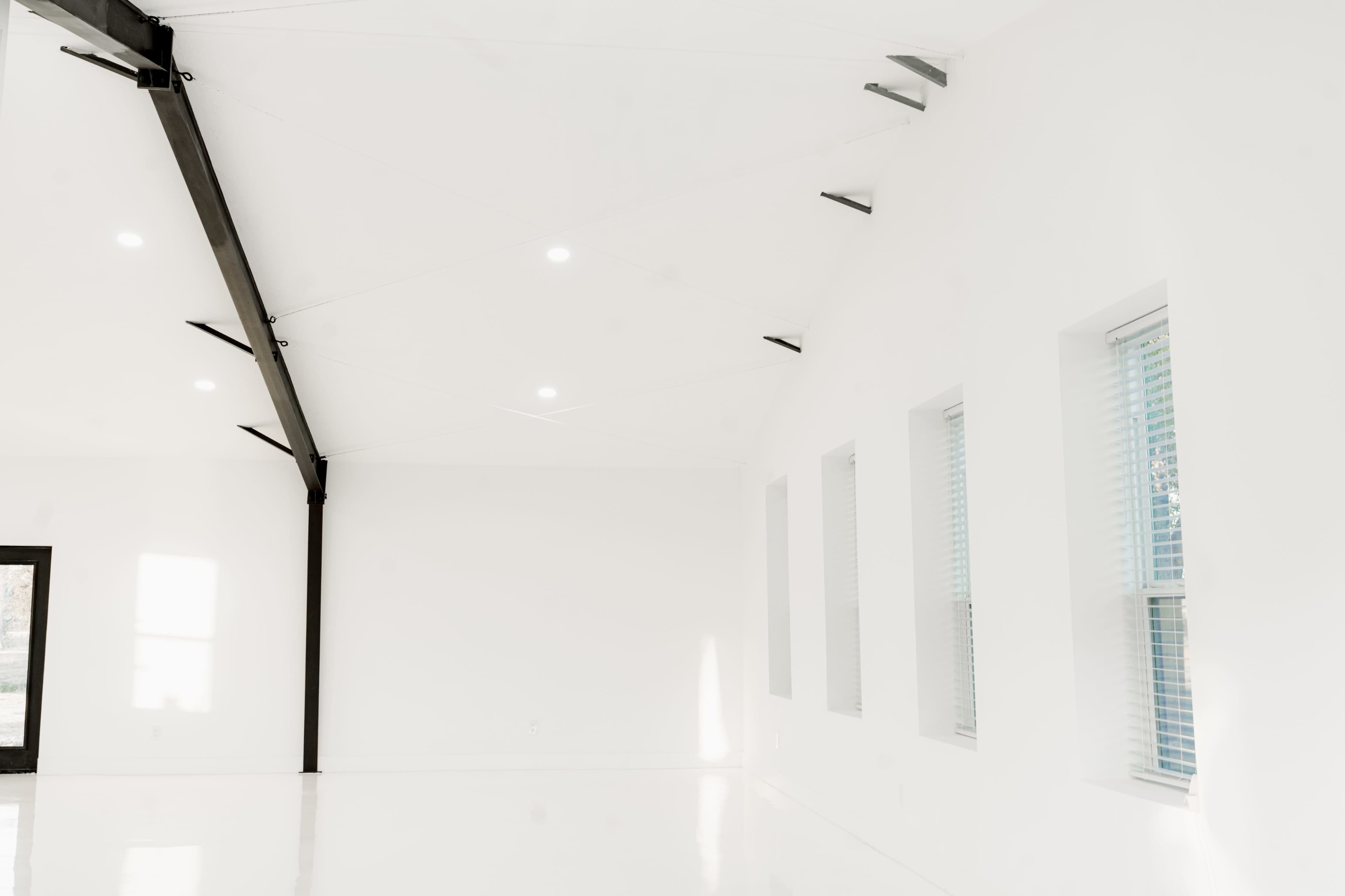 The image shows a spacious, minimalist room with white walls, large windows, and a high ceiling supported by black beams.