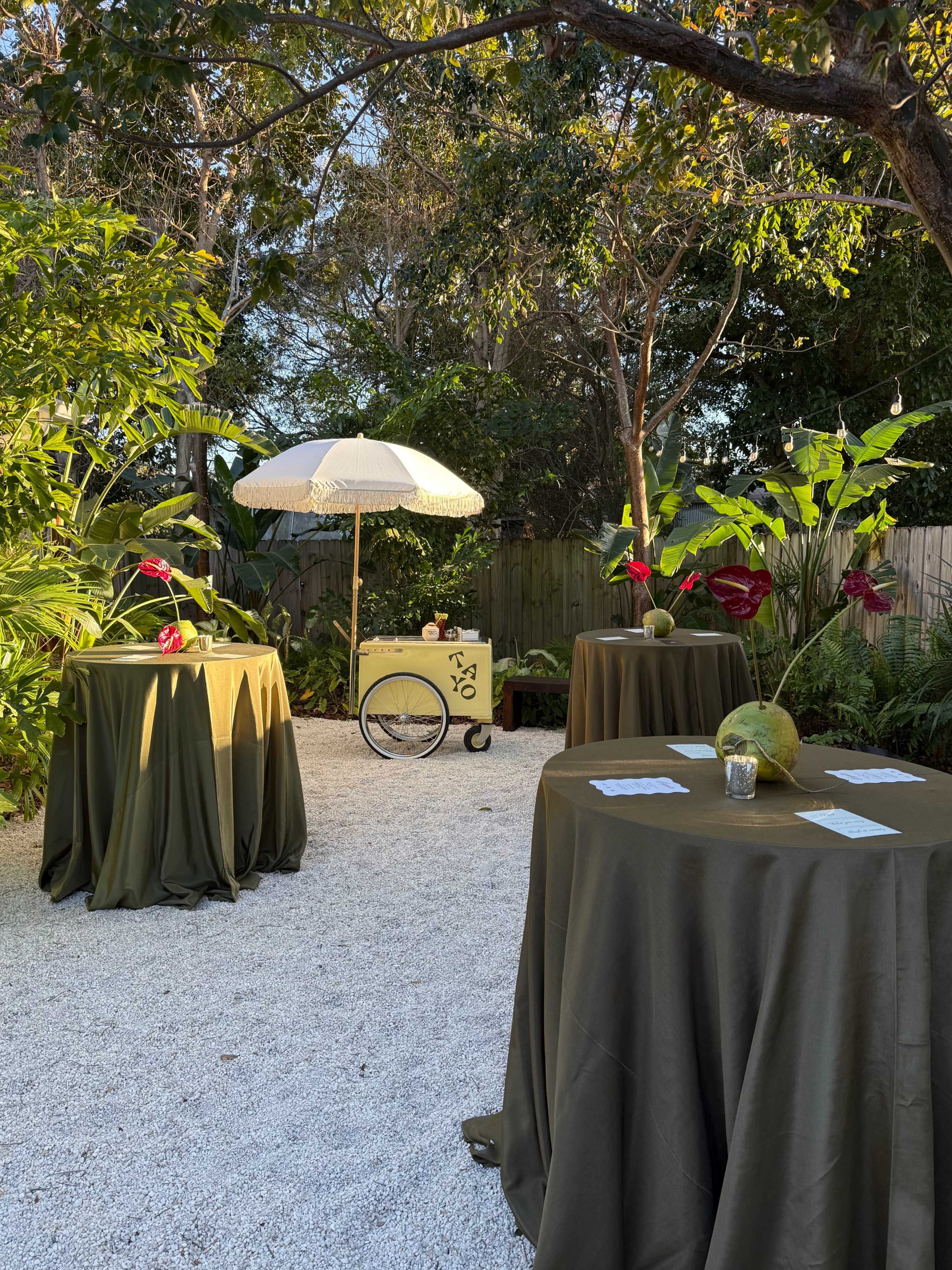 The scene features several round tables covered with olive green tablecloths arranged in a garden with tropical plants, an antique cart under a parasol, and decorative flowers.