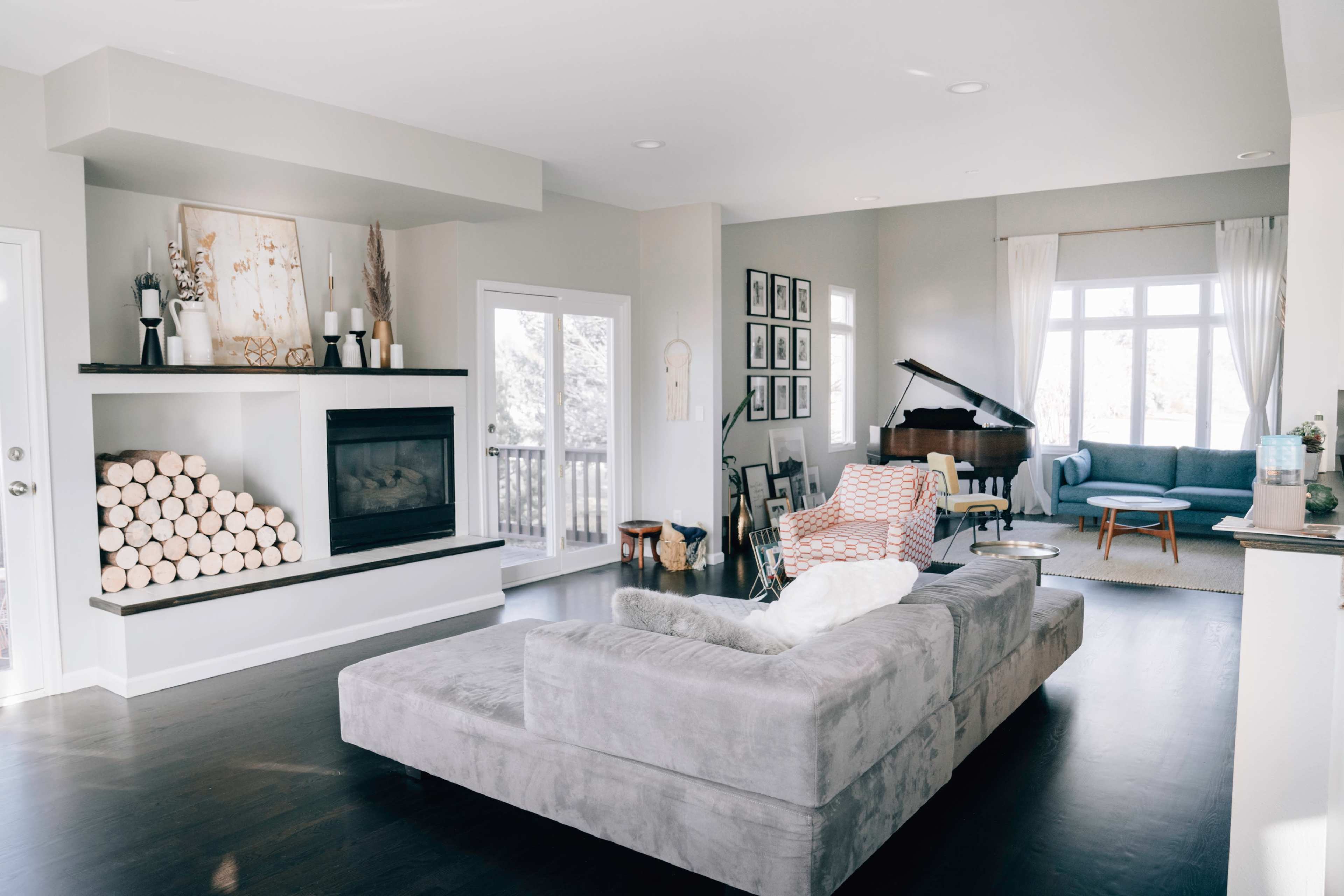 The image shows a modern living room with a gray sectional sofa, a fireplace with stacked logs, and a piano near a large window.
