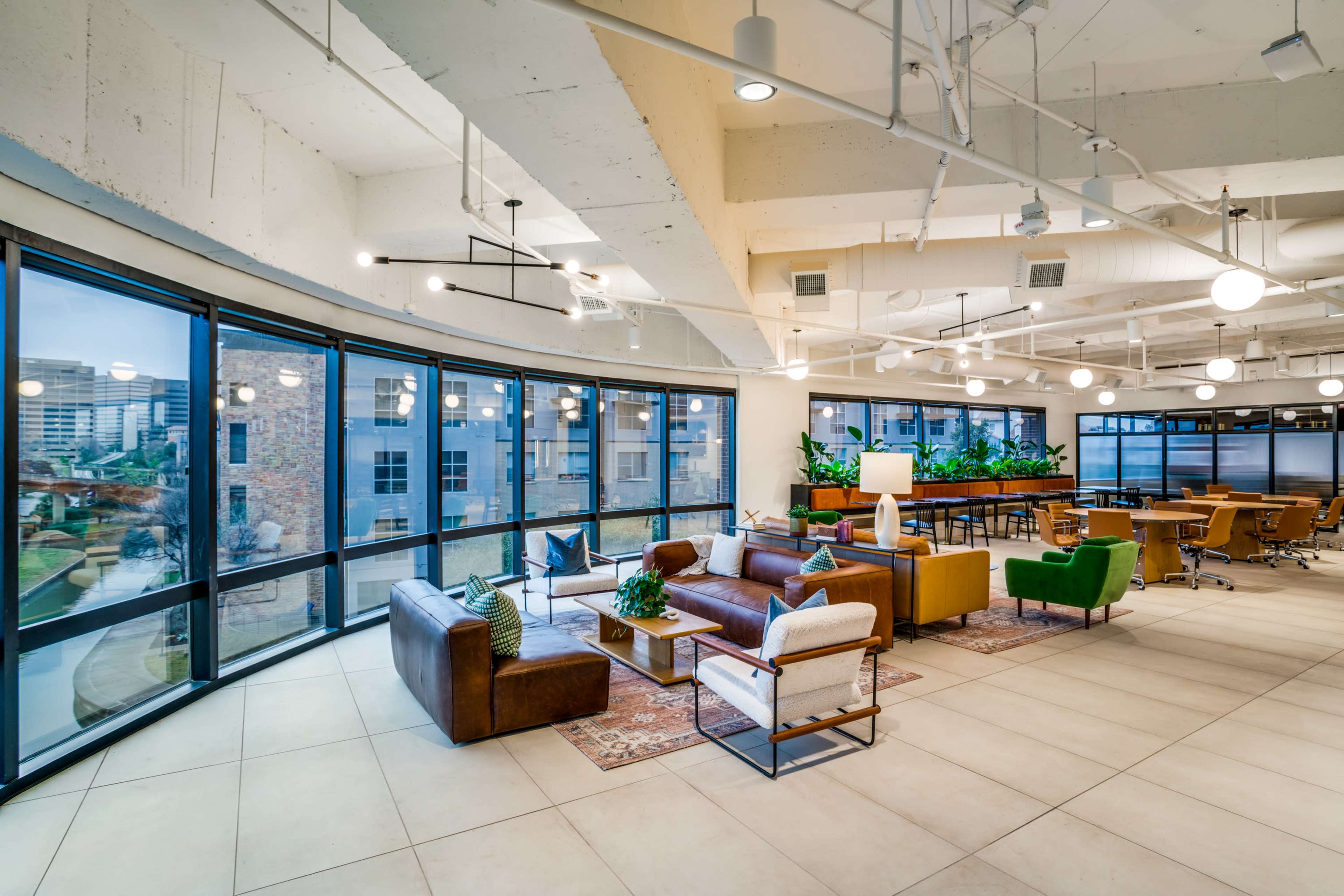 The image shows a modern office lounge area with large windows, featuring several seating arrangements, plants, and a workspace in the background.