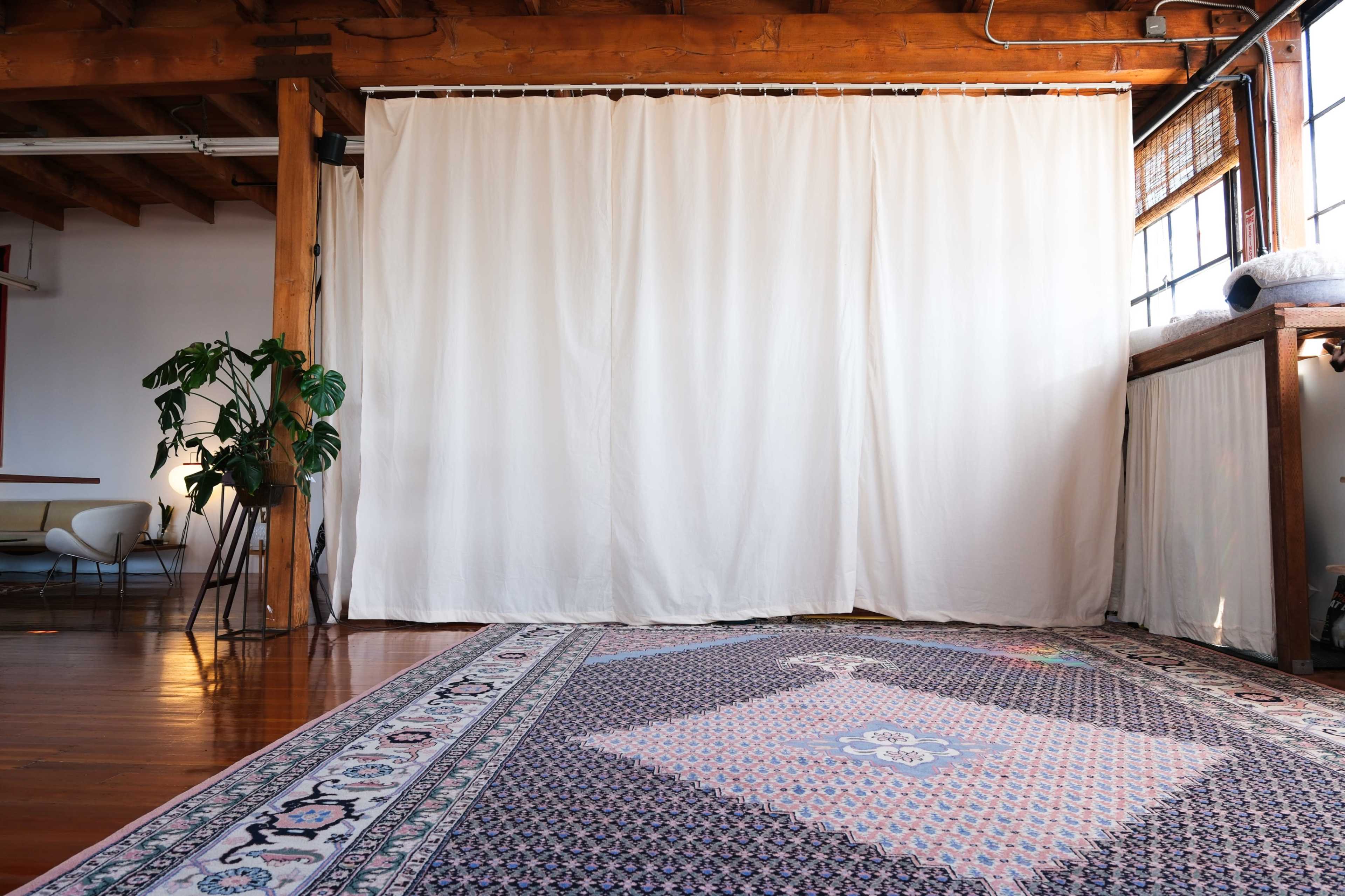 A spacious interior features a large white curtain, hardwood floors, and a patterned rug, complemented by a potted plant and a visible dining area.