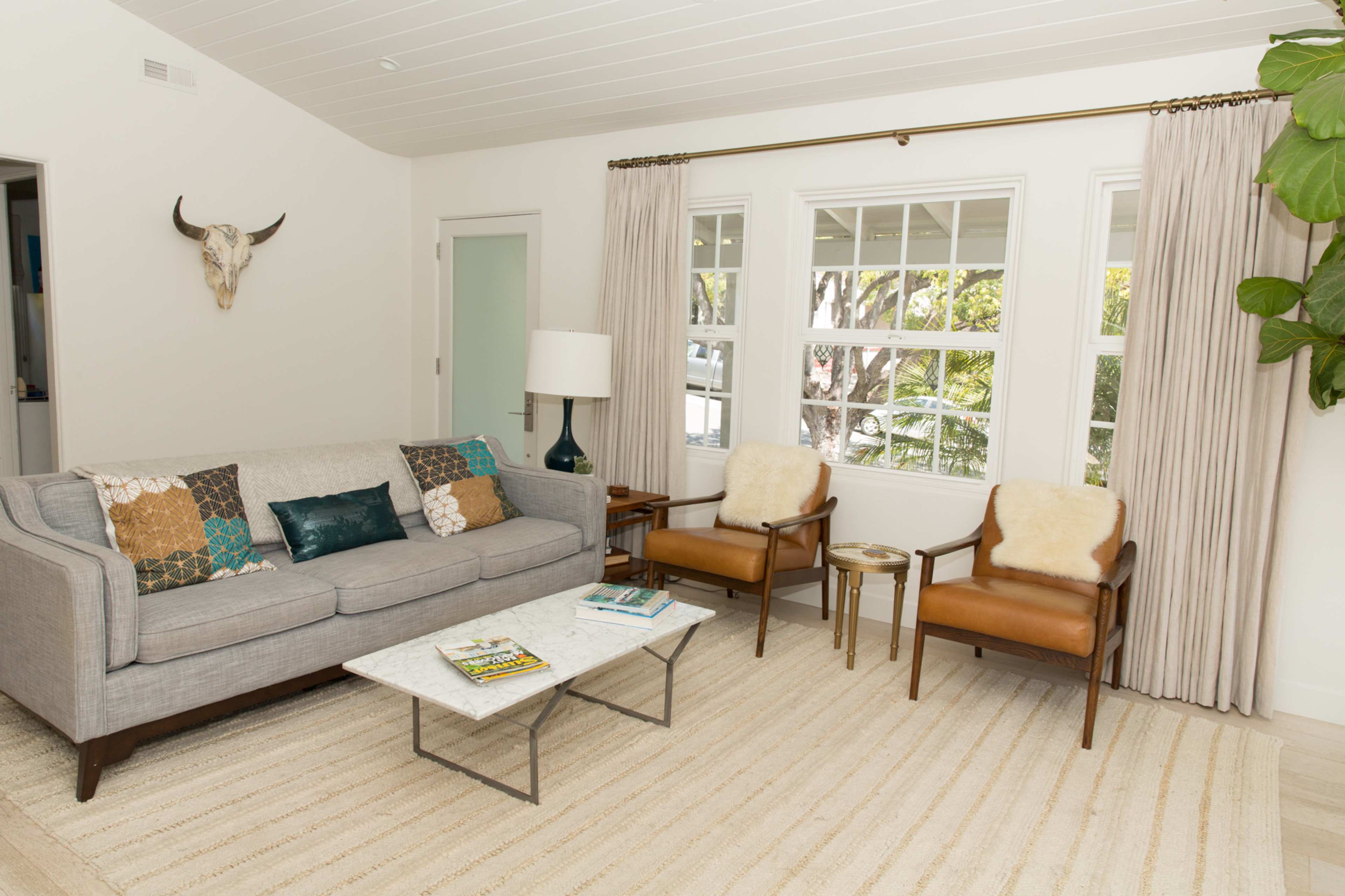 A modern living room features a gray sofa, two brown armchairs, a marble coffee table, and large windows with sheer curtains.