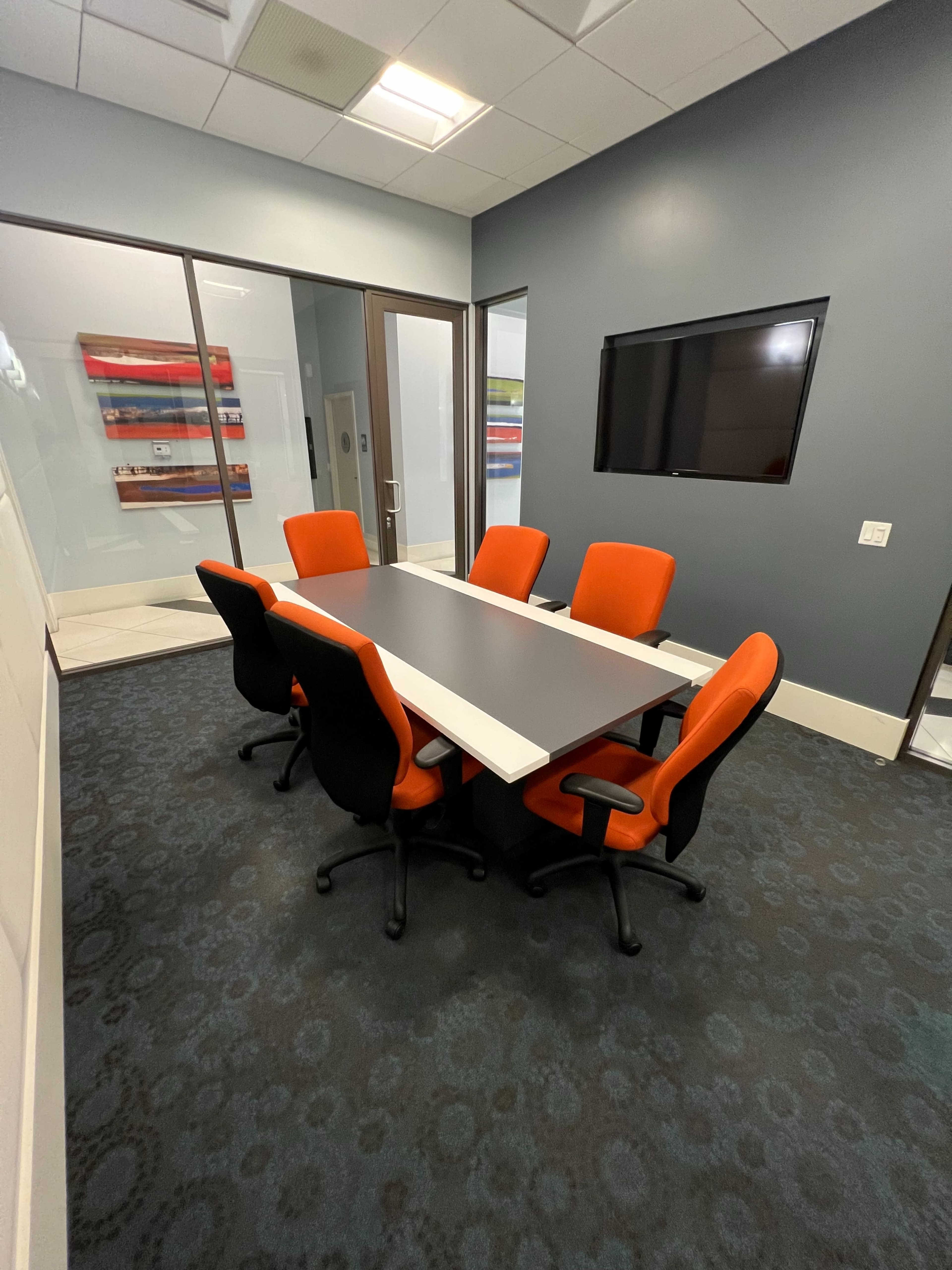 A modern conference room with a gray table surrounded by six orange and black chairs, featuring a wall-mounted TV and glass partition walls.