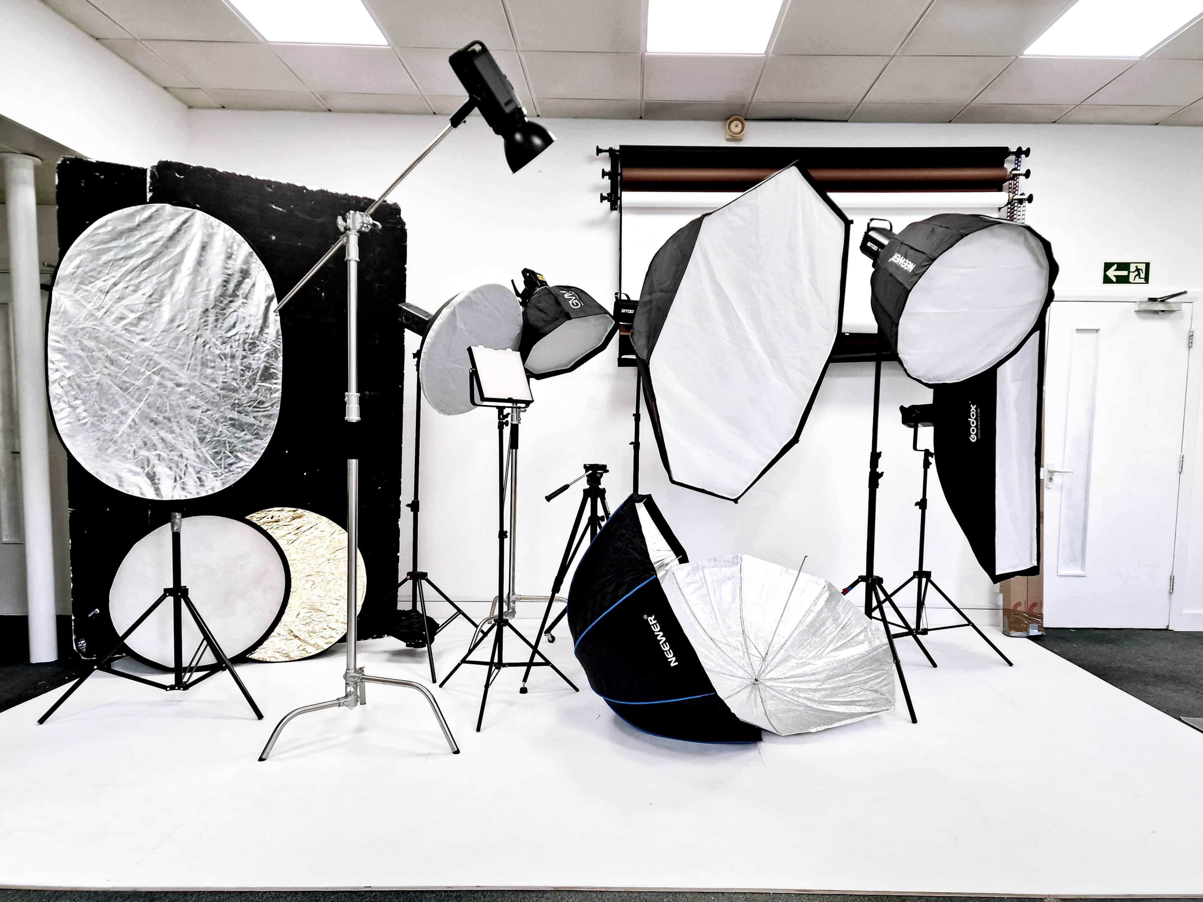A photography studio is set up with various lighting equipment, including softboxes, reflectors, and light stands arranged on a white backdrop.