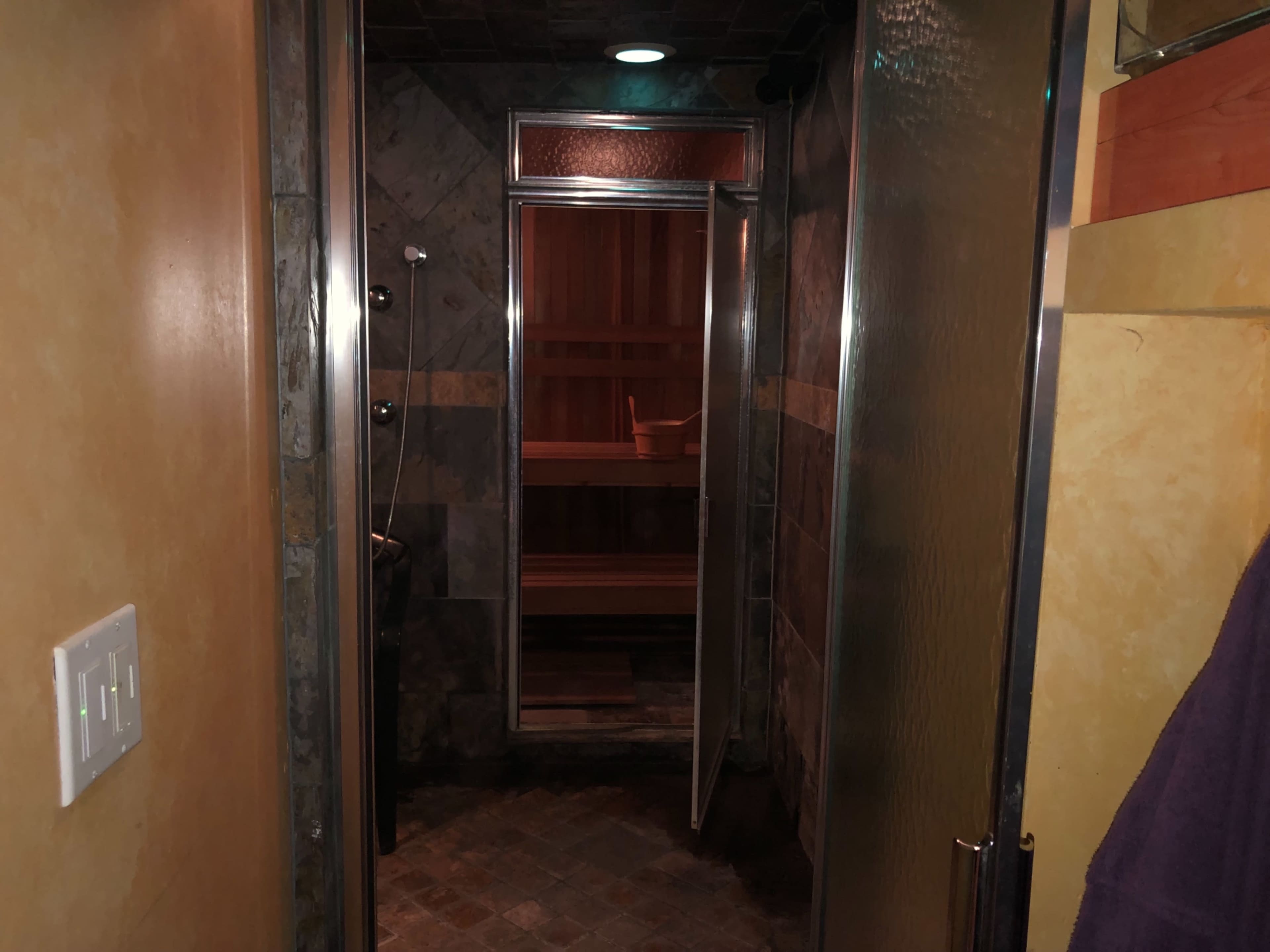 A tiled bathroom with a glass shower door and a wooden sauna visible in the background.