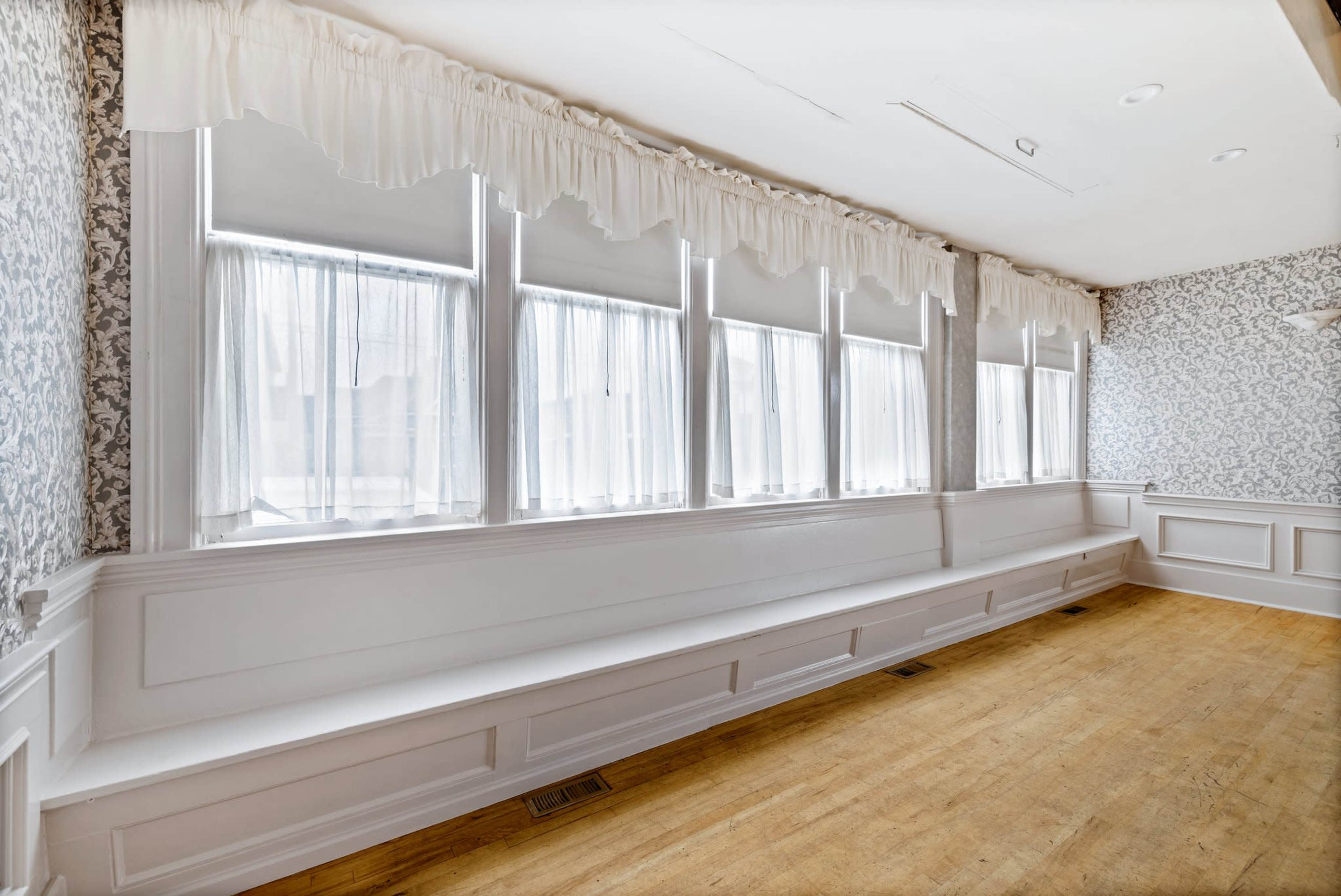 A brightly lit room features large windows with white curtains and patterned wallpaper.