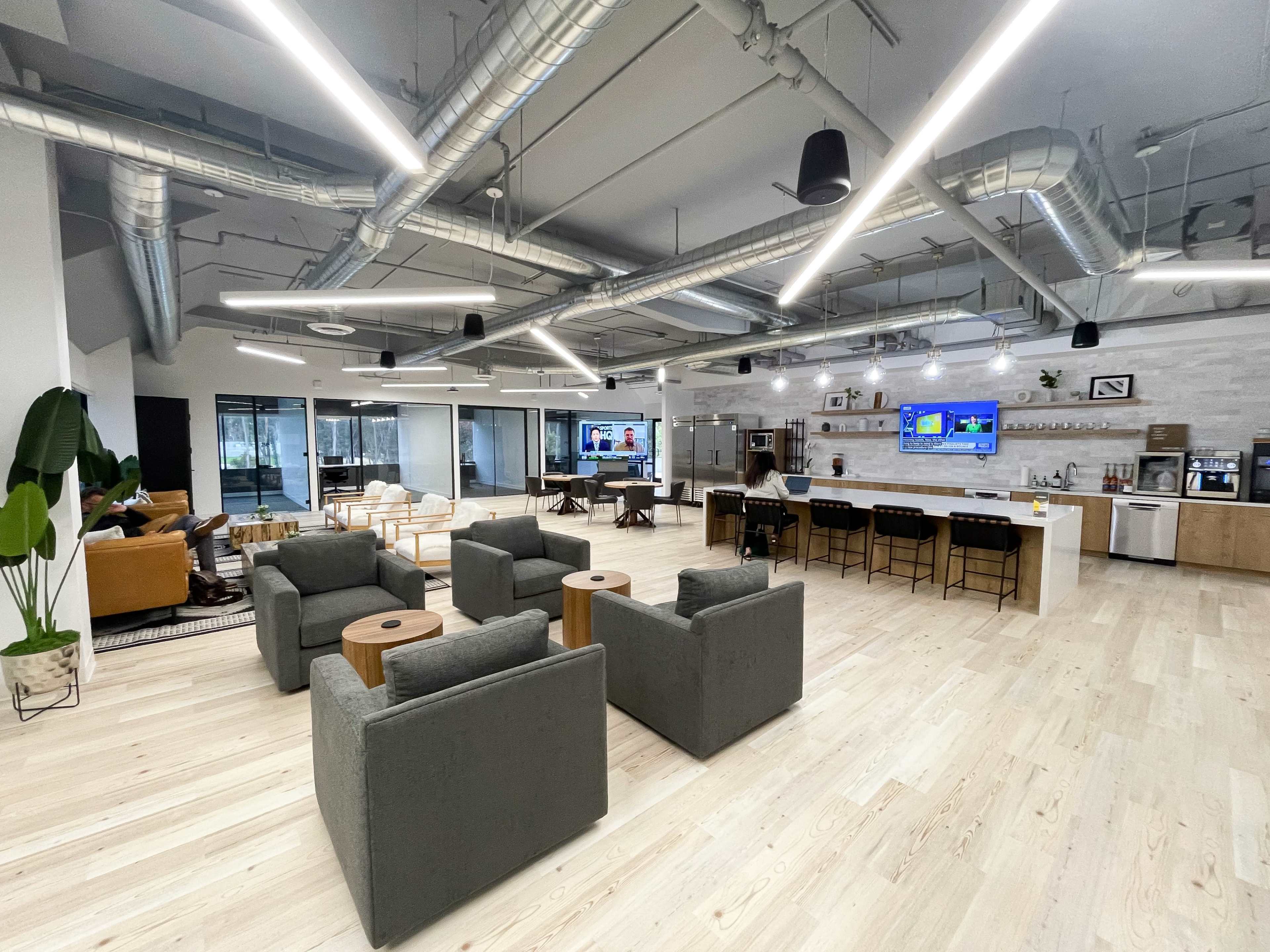 A modern office space with a lounge area featuring gray sofas and wooden tables, a kitchen area, and large windows letting in natural light.