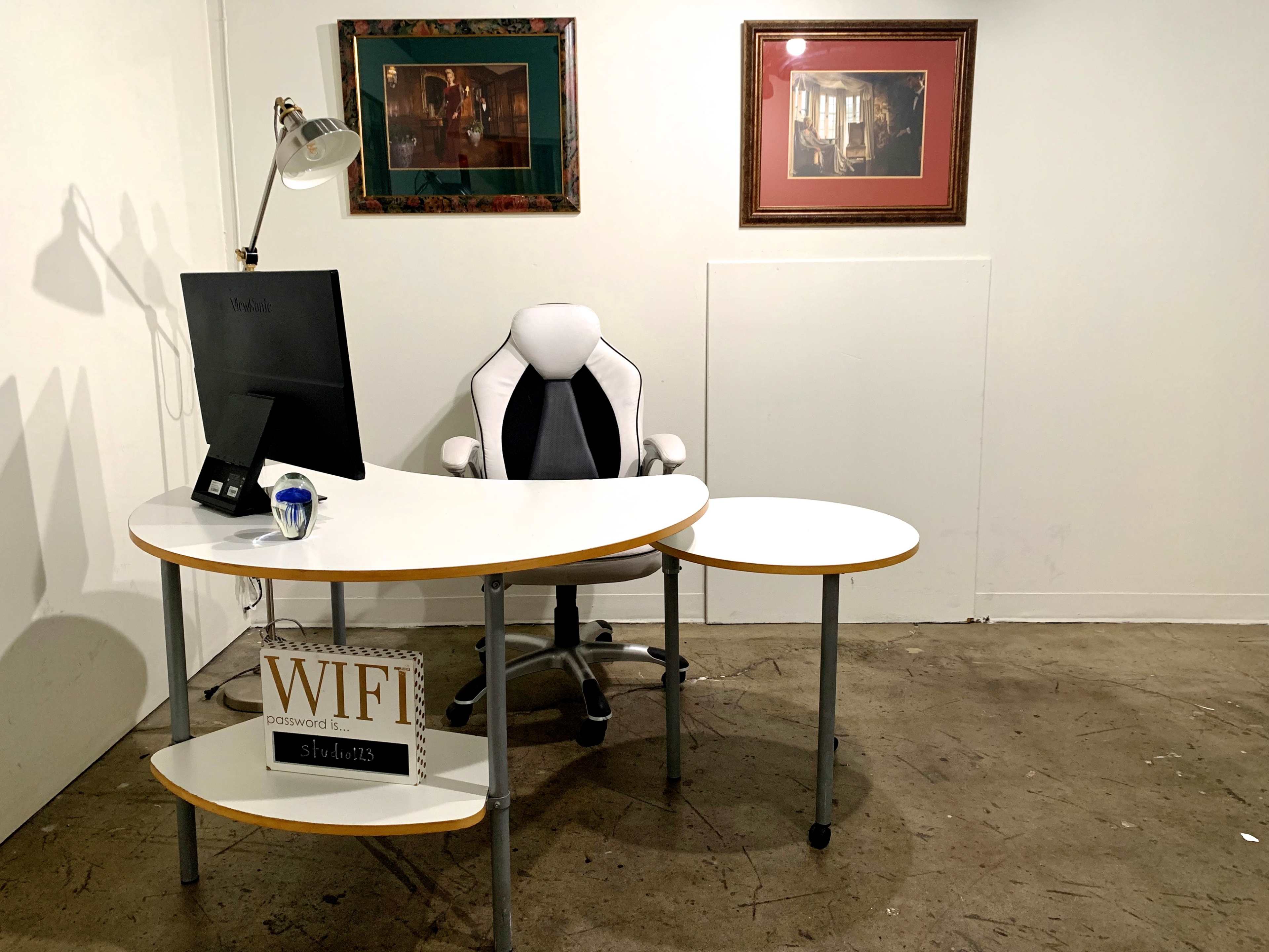 A modern office space features a white desk with a computer monitor and a rolling office chair, accompanied by framed artwork on the wall.