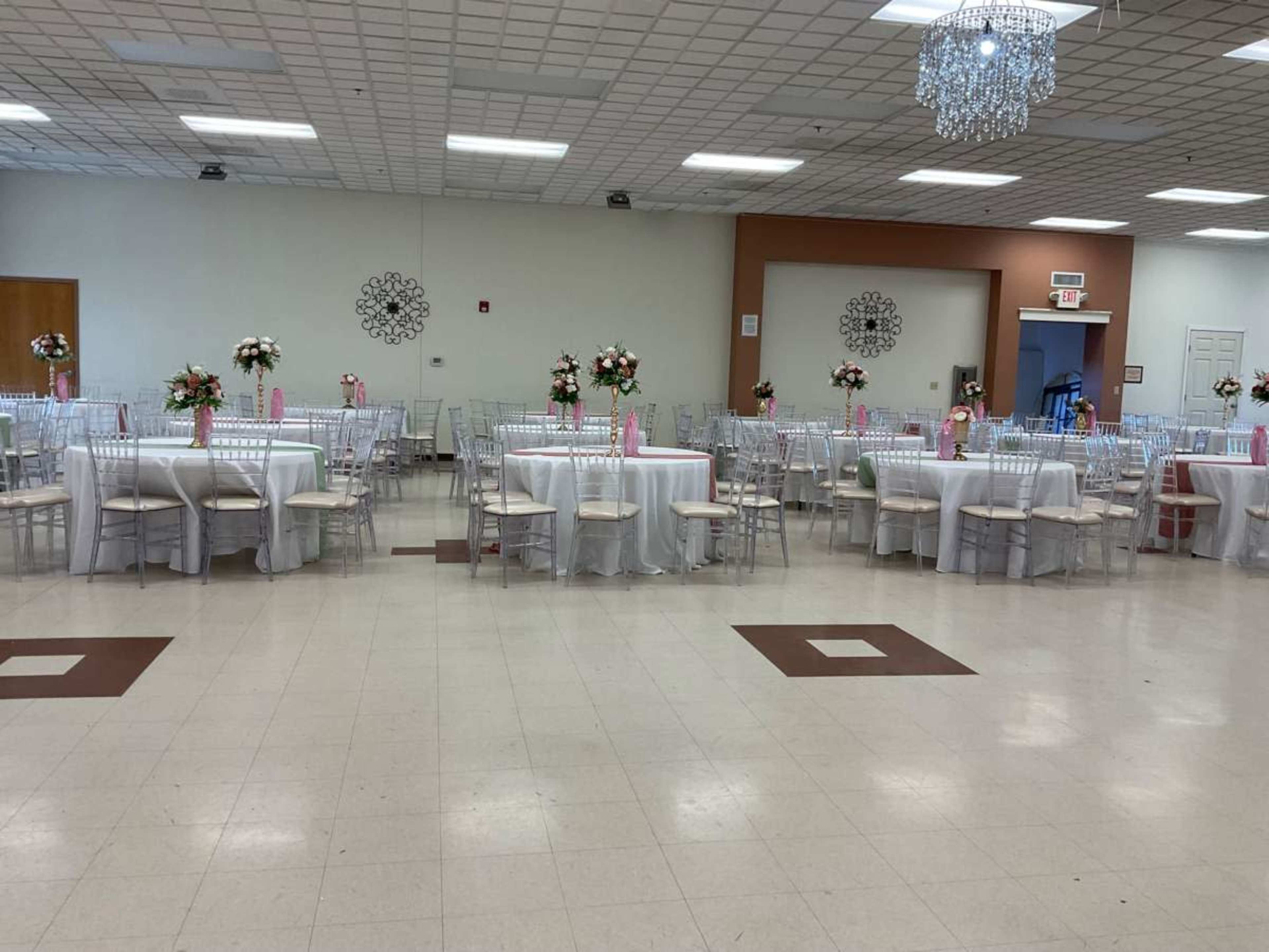 The image depicts an elegant event space with round dining tables covered in white tablecloths, each adorned with floral centerpieces and set with clear ghost chairs.