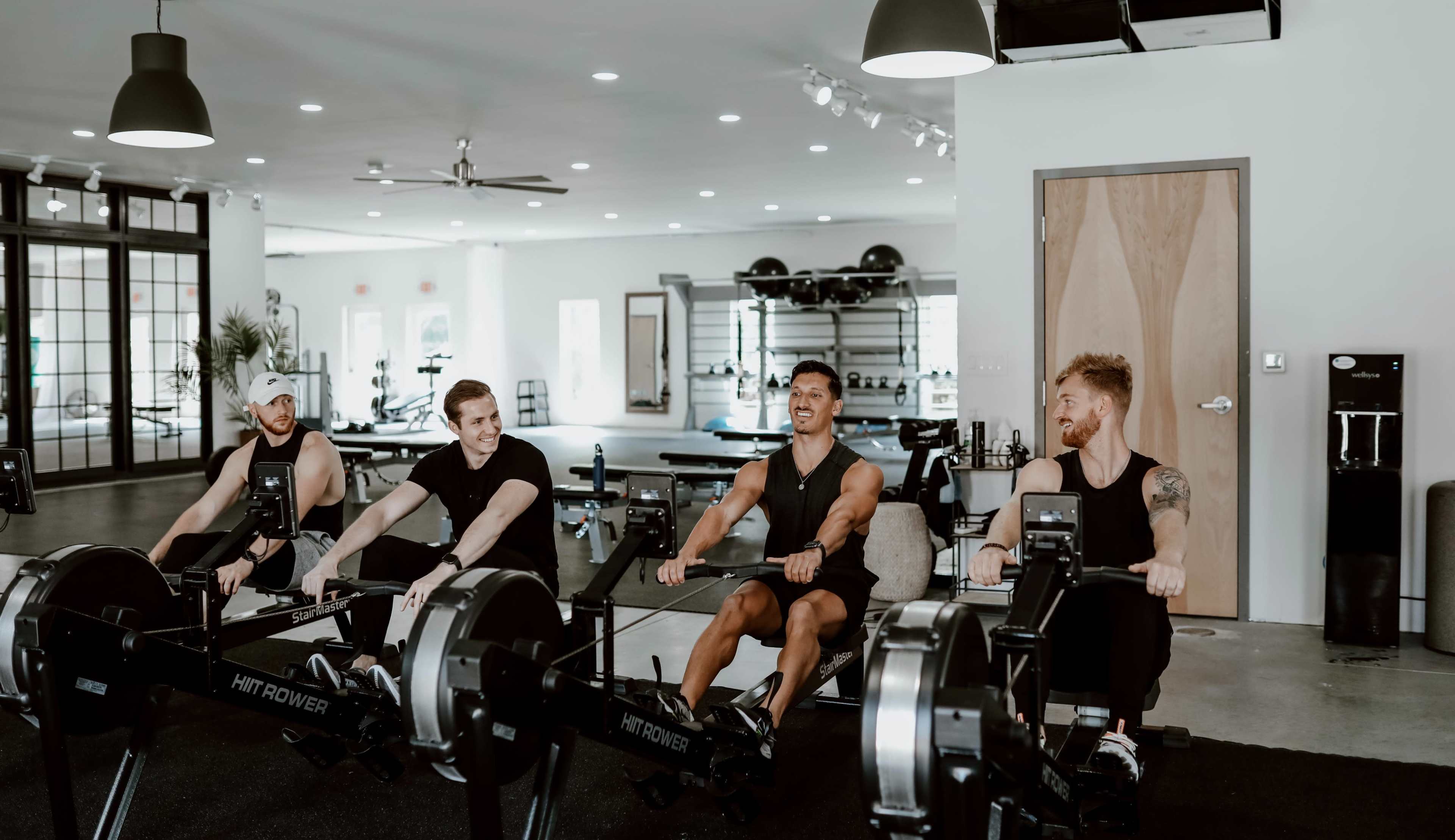 Four men are exercising on rowing machines in a modern gym setting.