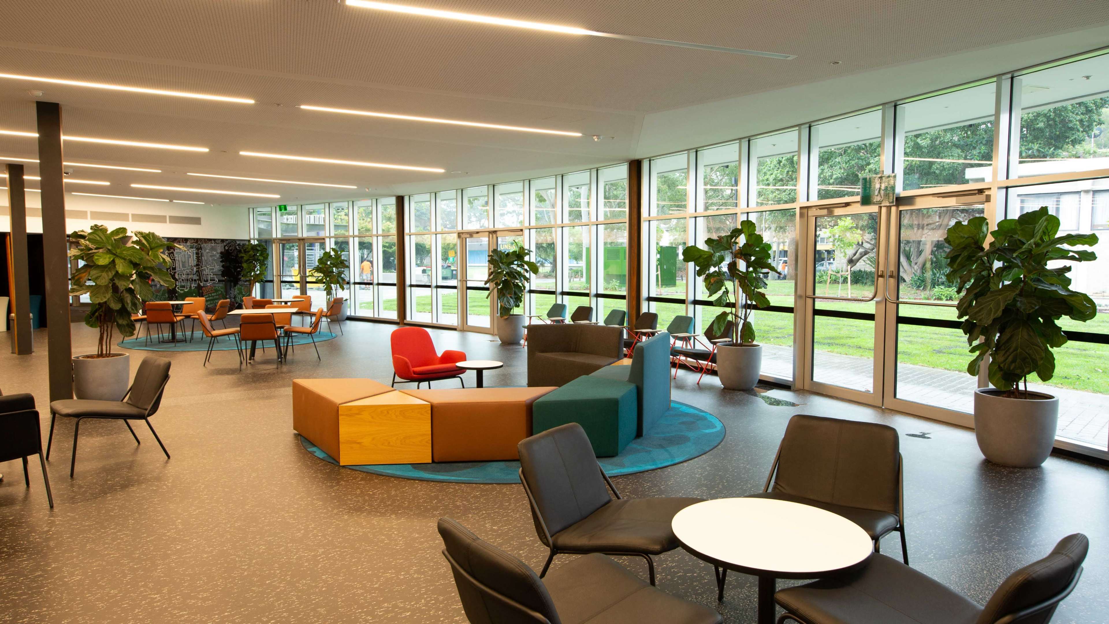A modern lounge area with seating arrangements, including chairs and tables, surrounded by large windows and indoor plants.