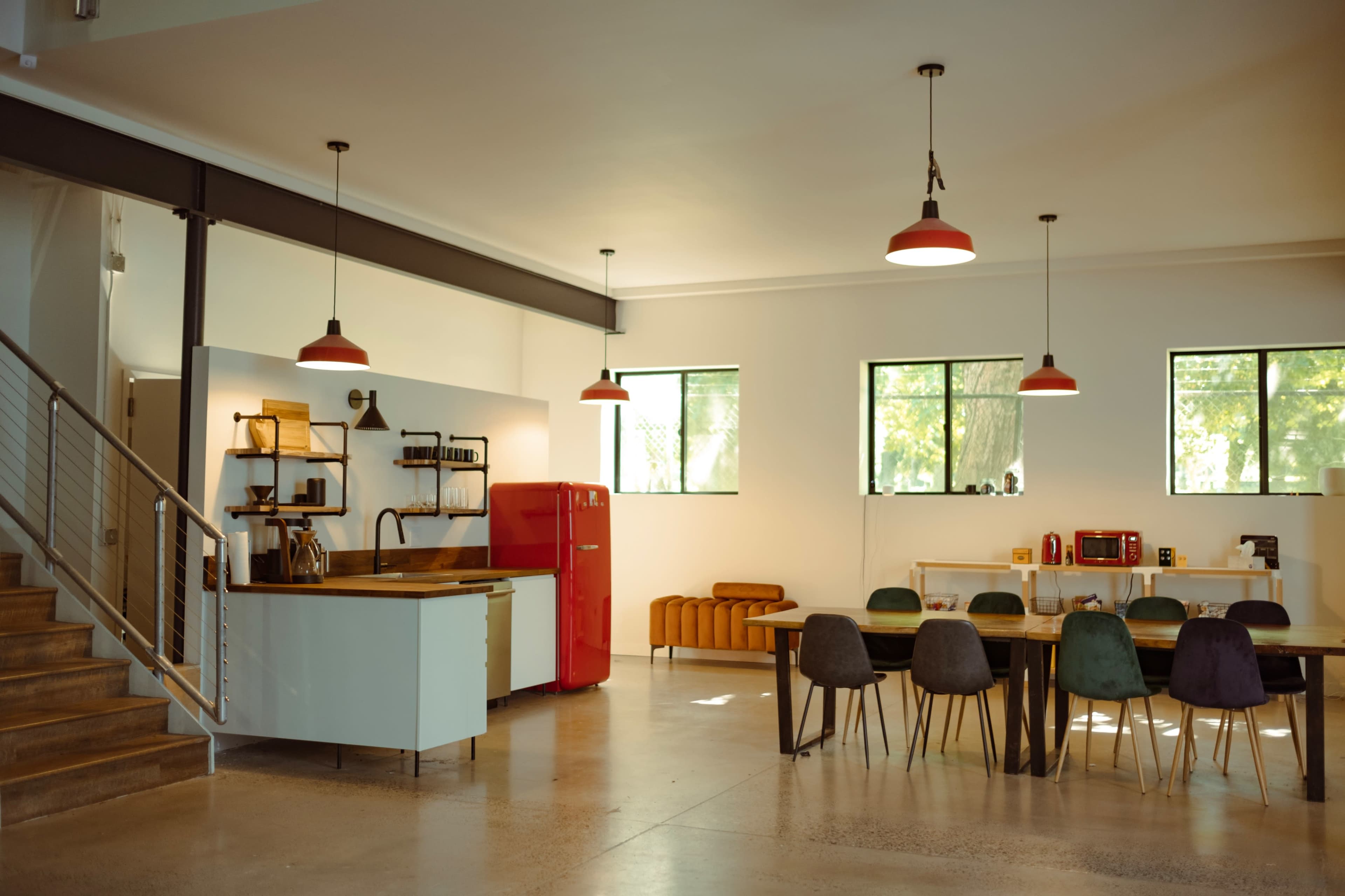 A spacious modern kitchen and dining area with a red refrigerator, wooden dining table, and large windows allowing natural light to enter.
