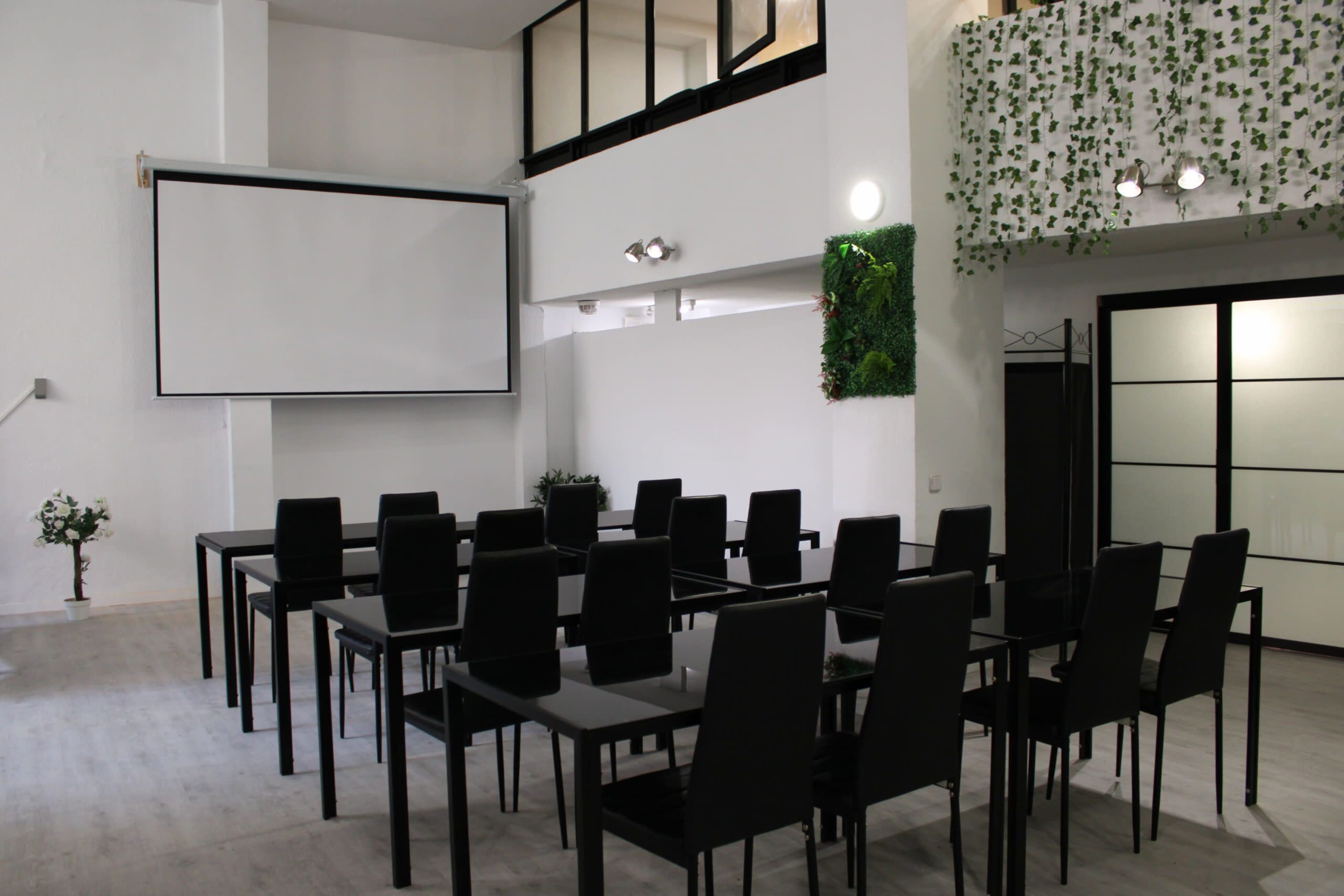 The image shows a modern meeting room set up with rows of black chairs and tables, featuring a large screen and a decorative plant wall.