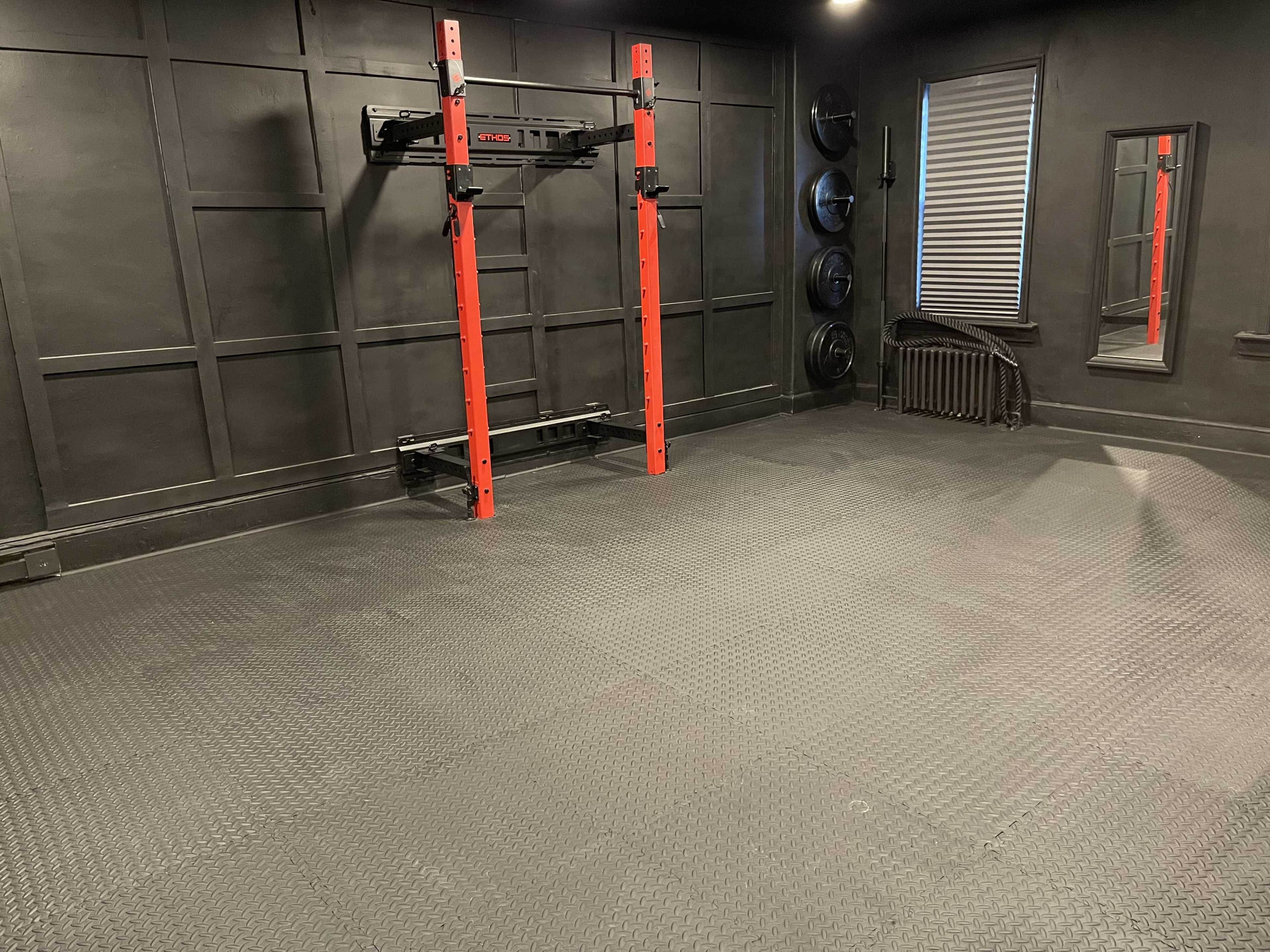 A black-painted gym interior with a squat rack, weight plates, a mirror, and rubber flooring.