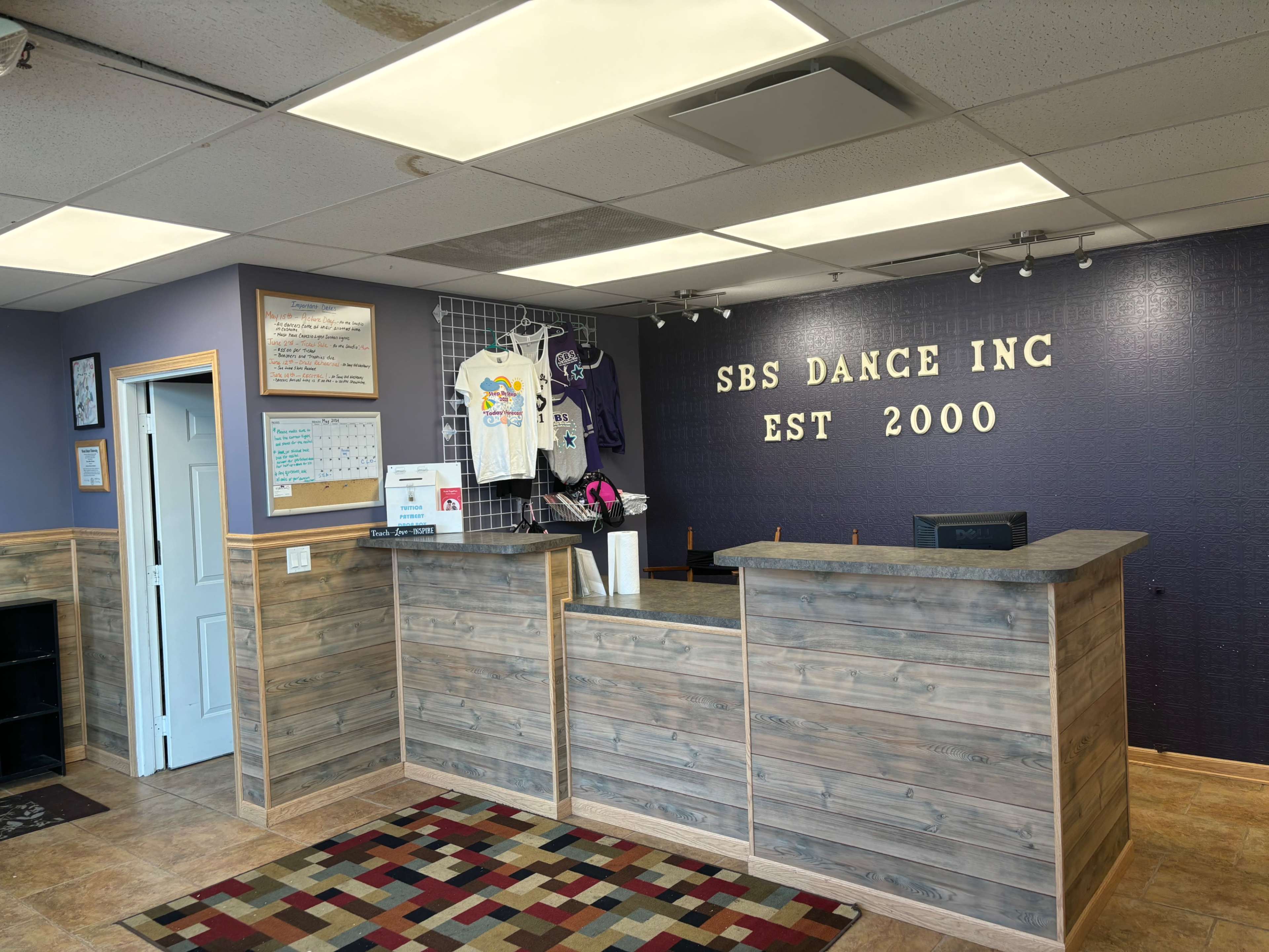 The interior of a dance studio reception area with a wooden front desk and promotional items displayed on a wall grid.