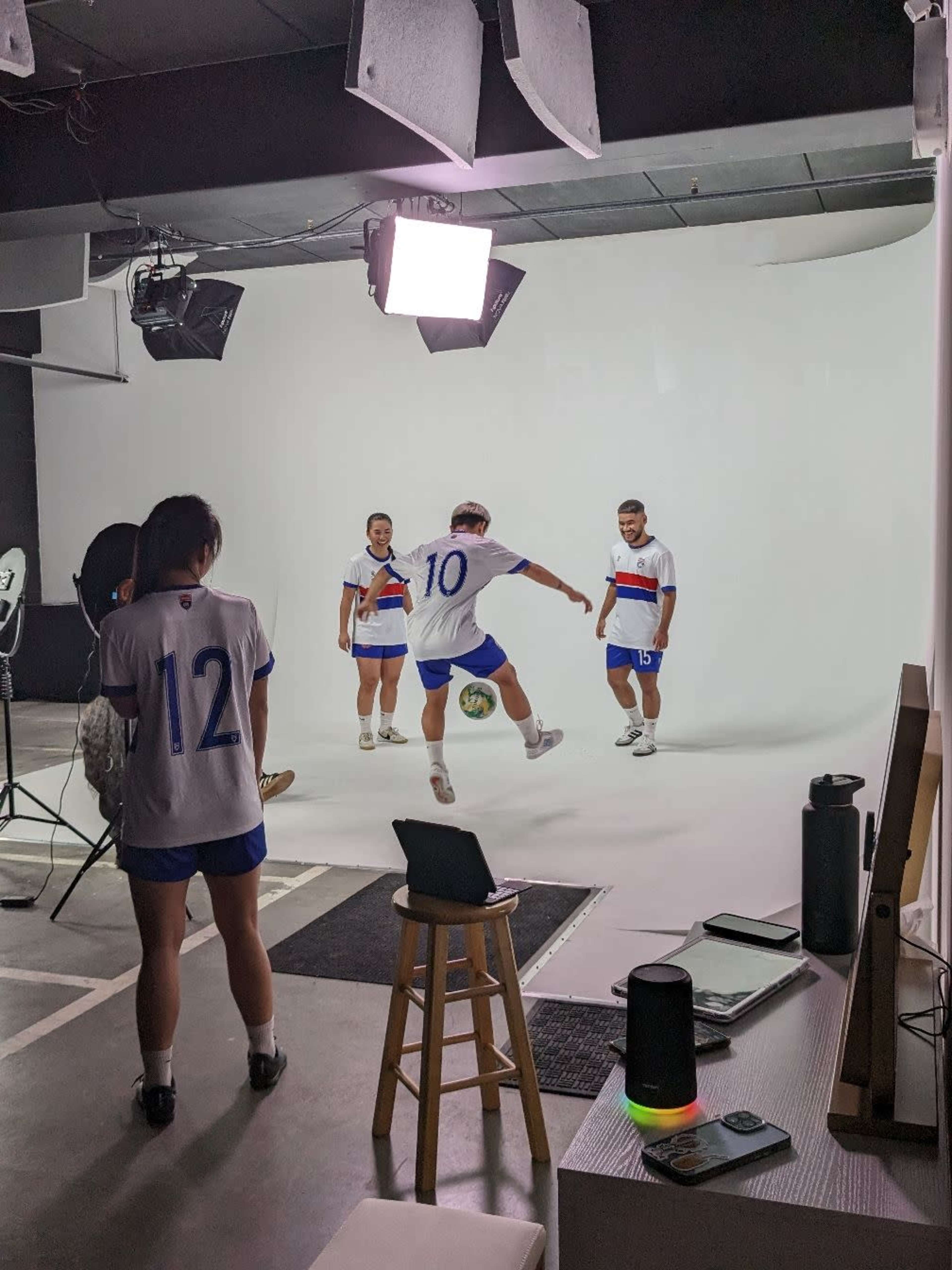 Three soccer players in matching white jerseys pose for a photoshoot against a plain white background, while one person practices juggling a soccer ball.