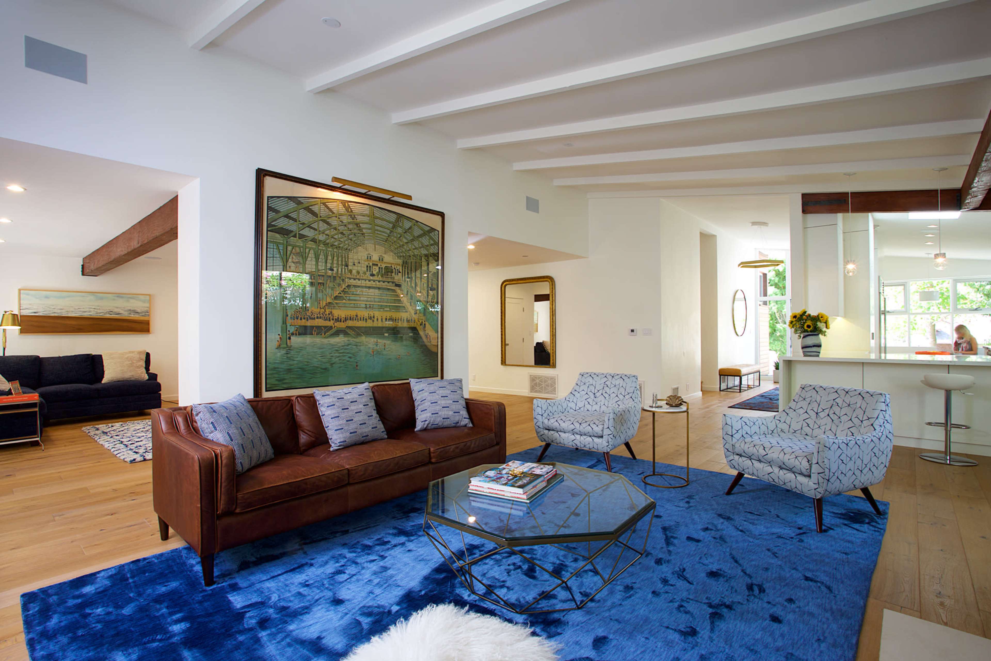 A spacious living room features a brown leather sofa, patterned chairs, a glass coffee table on a blue rug, and a large artwork depicting an indoor pool.