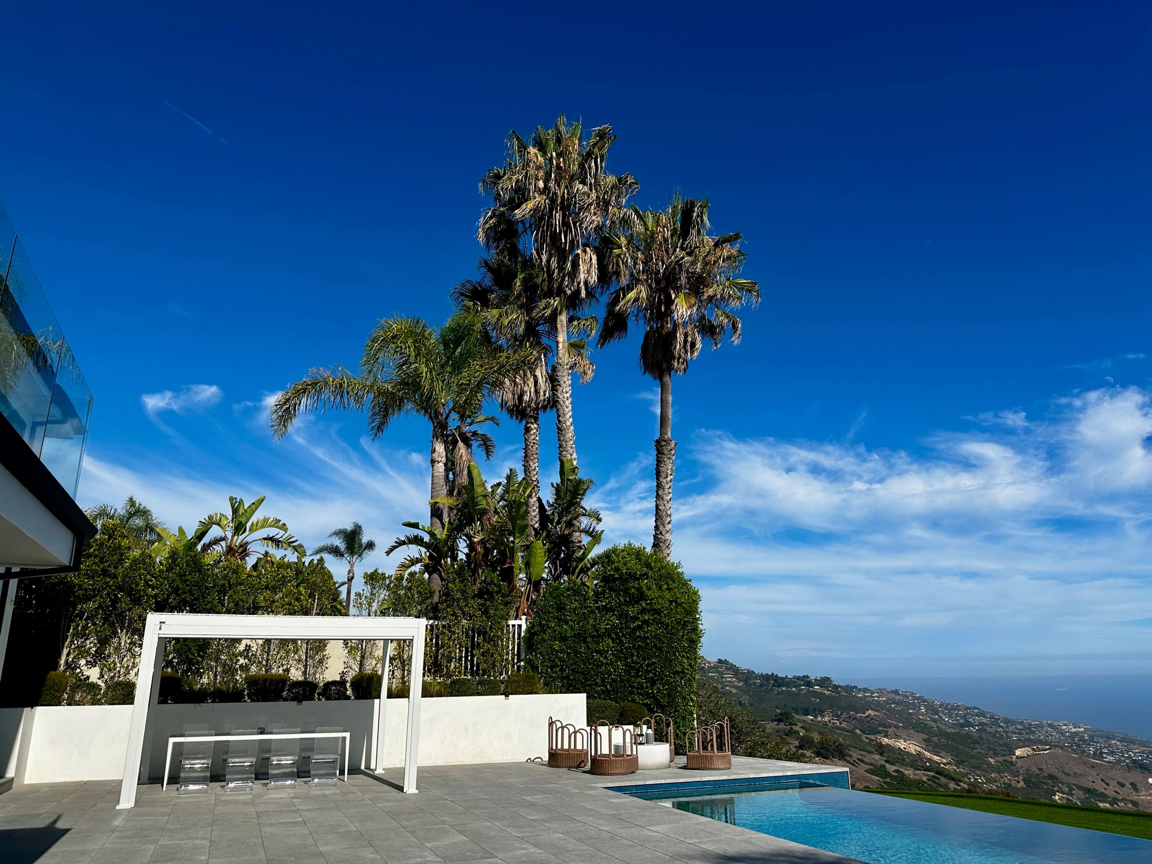 The image shows a modern luxury home with a pool, surrounded by palm trees and a coastal view under a clear blue sky.