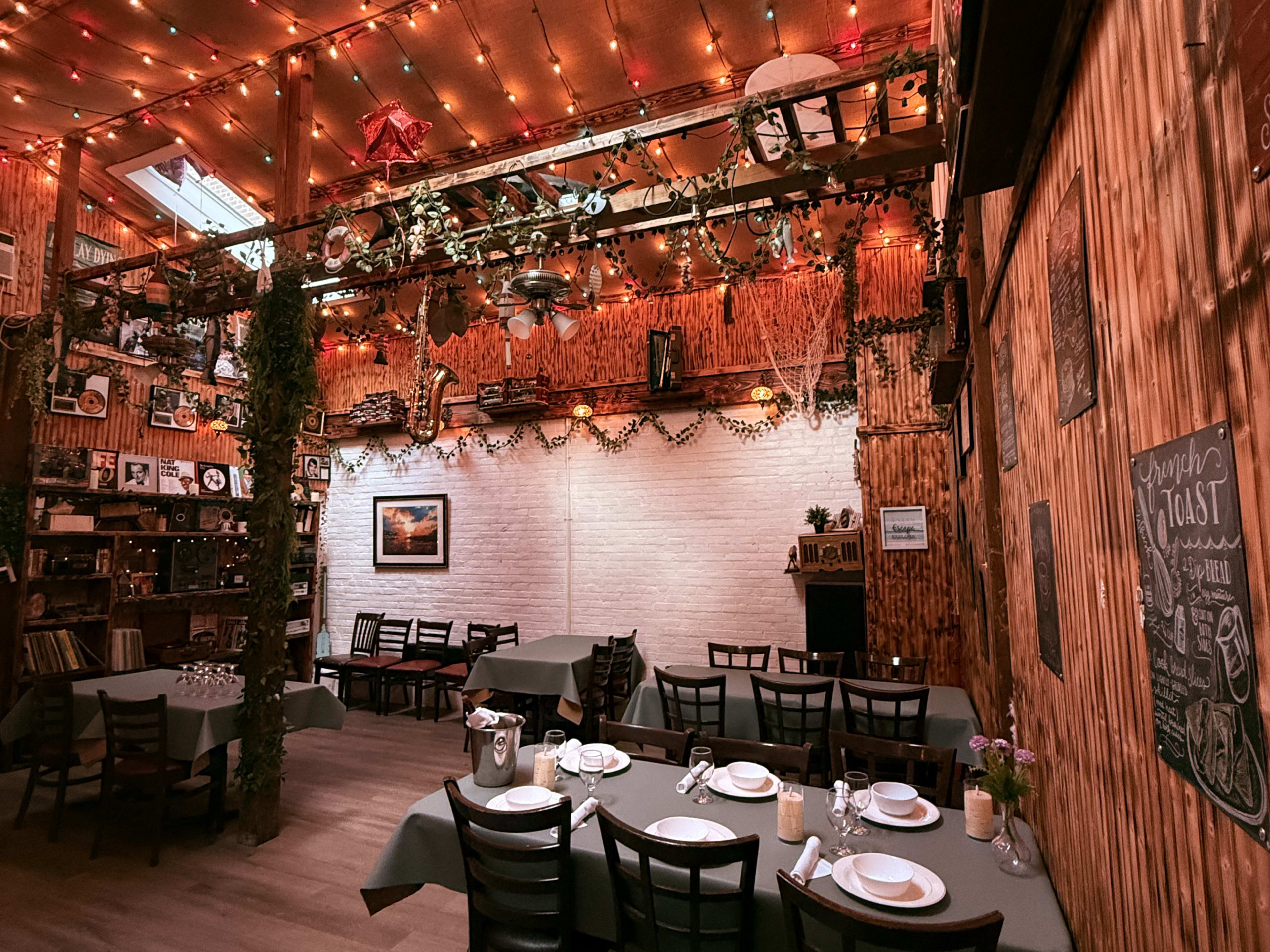 The image shows a warm, rustic restaurant interior with wooden walls, tables set with dishware, and decorative lights hanging above.