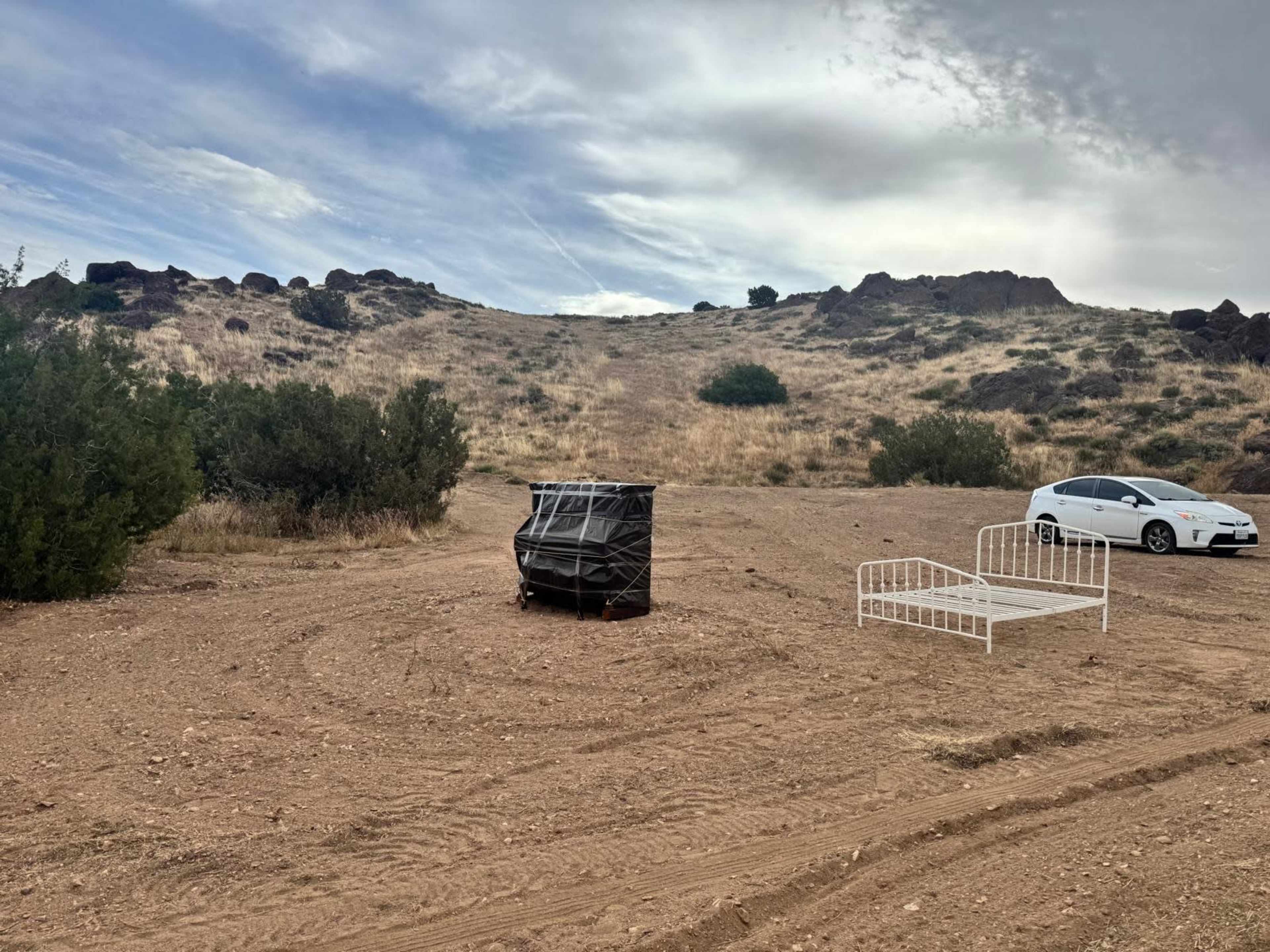 A black, wrapped object sits next to a white bed frame in a sandy area surrounded by sparse vegetation and rocky hills.