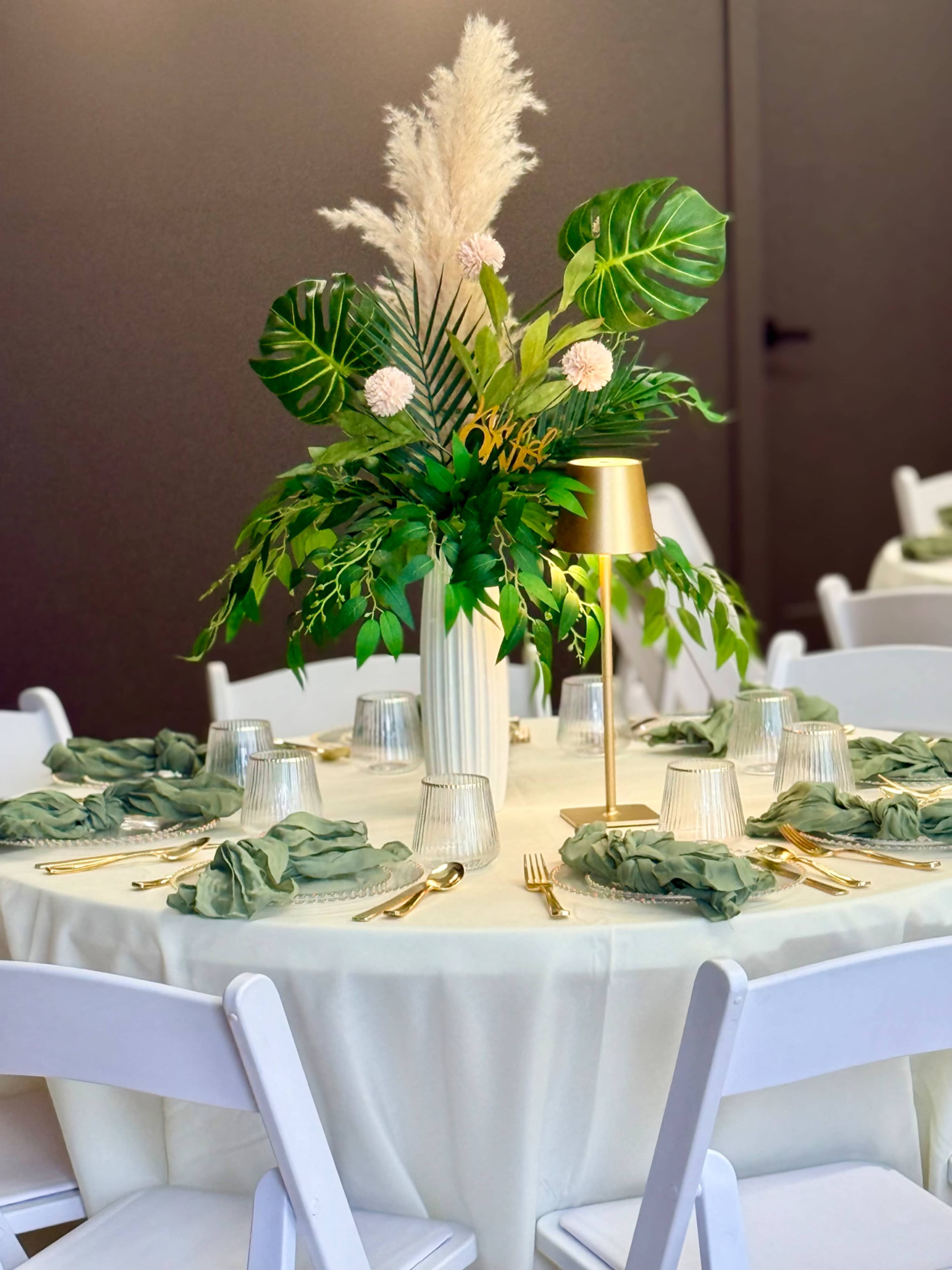 A elegantly set dining table features a tall floral centerpiece with pampas grass and green foliage, surrounded by neatly arranged place settings with green napkins and gold utensils.
