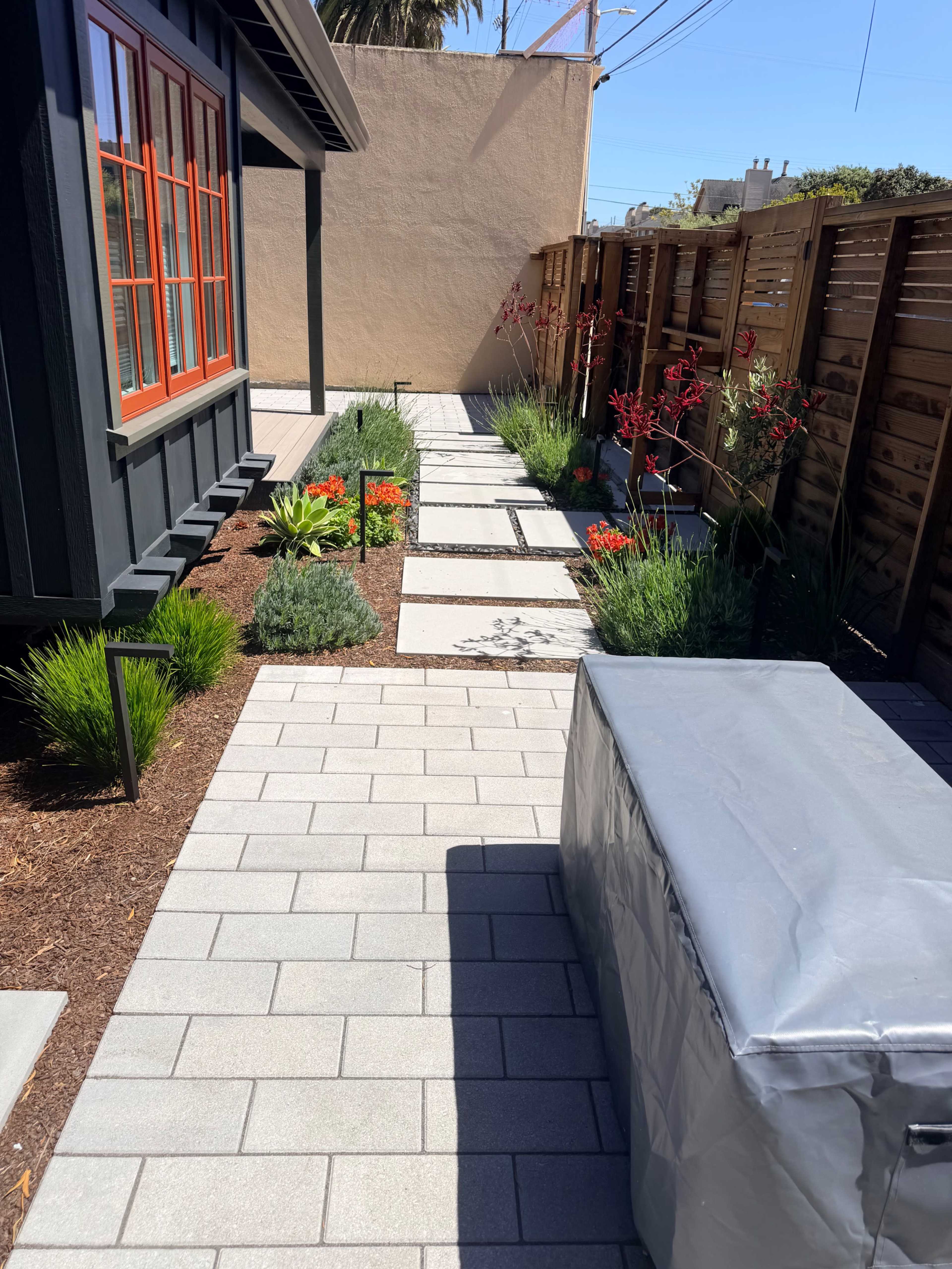 The image shows a paved pathway lined with various plants and flowers, leading to a modern black building with bright red windows.
