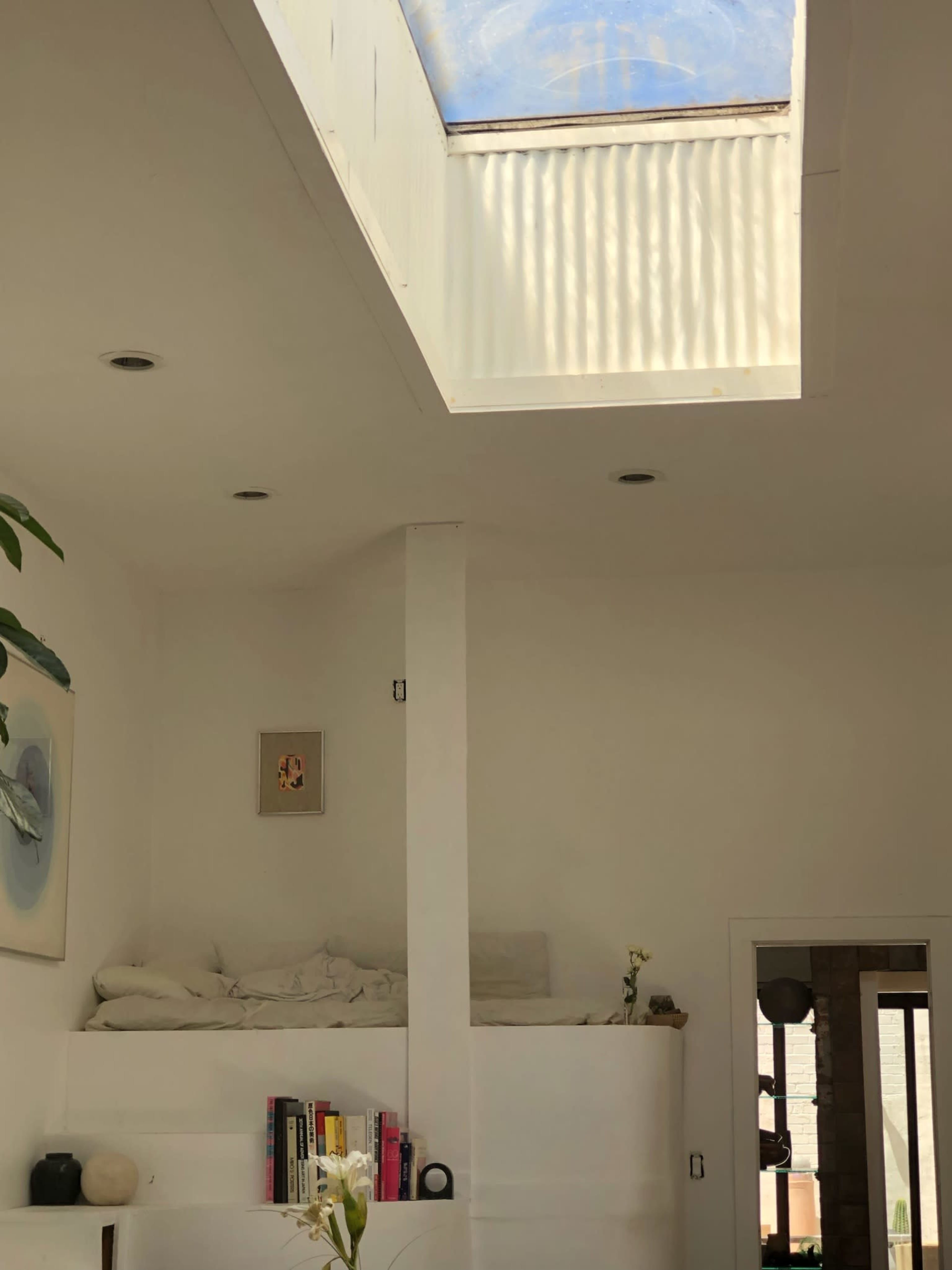 The image shows a sparsely furnished white interior with a skylight above a raised sleeping area and a small bookshelf.