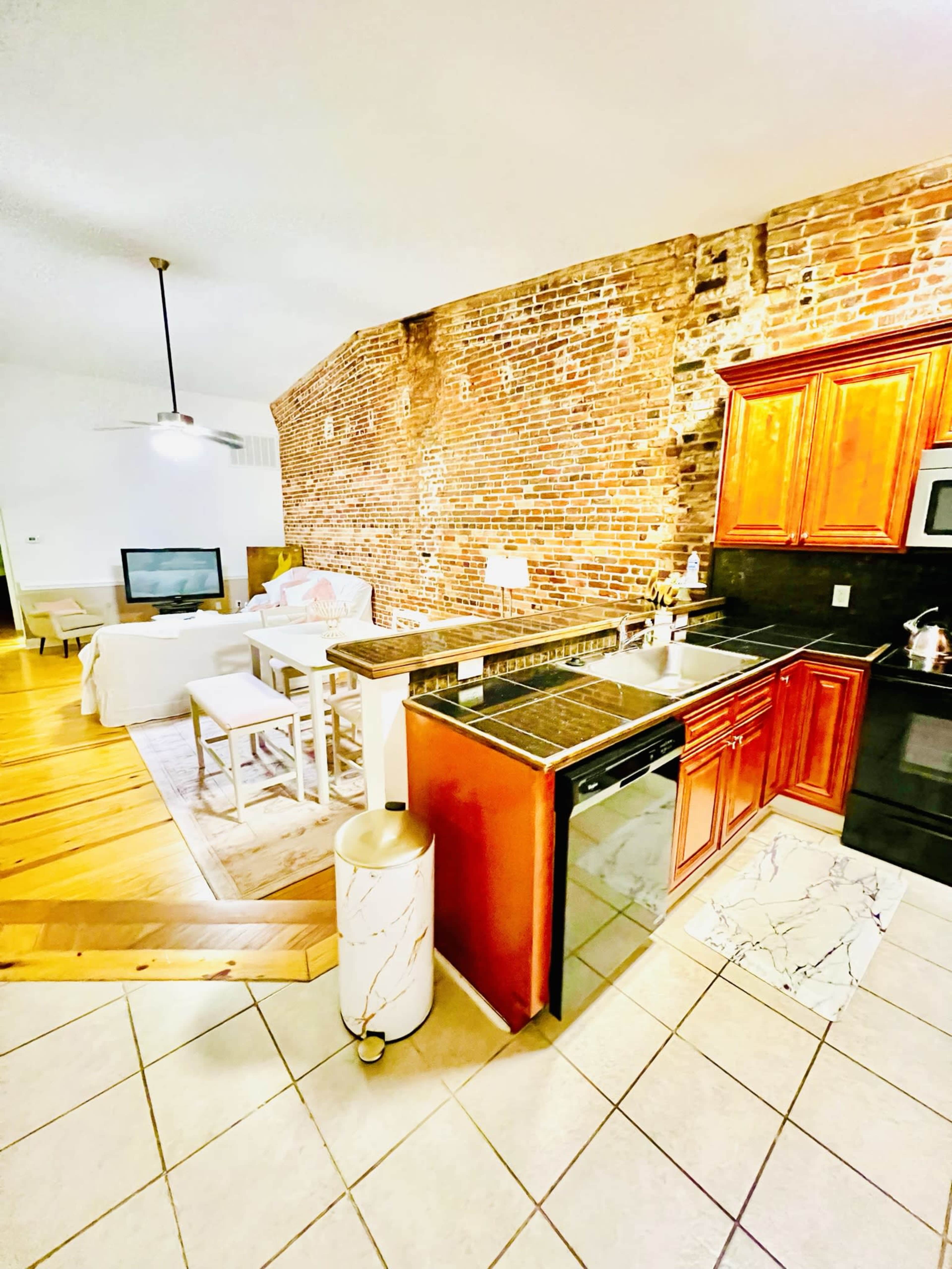 A kitchen with a brick wall, featuring dark cabinetry, a dishwasher, and a view of a living area with a television.
