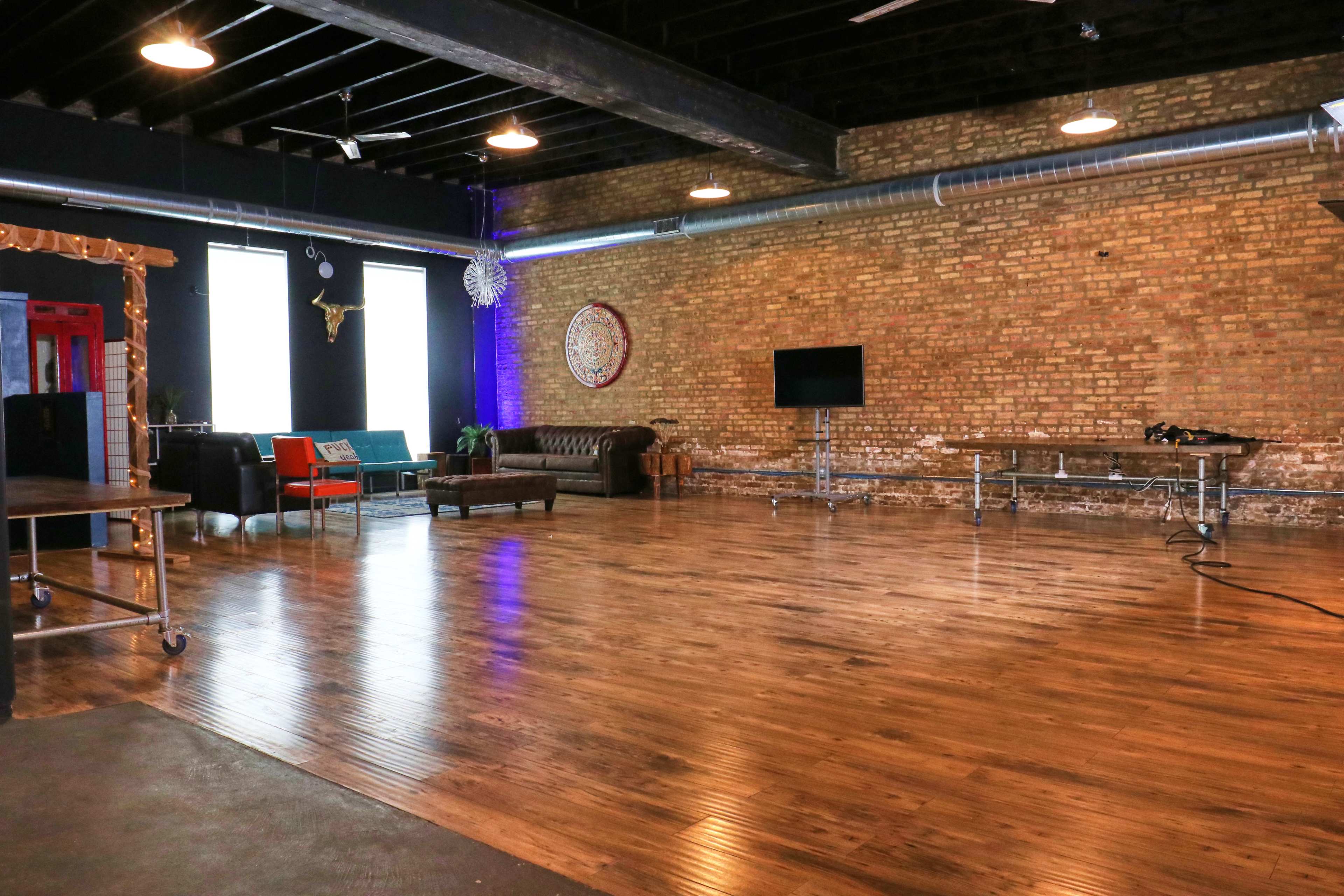 The image shows a spacious, industrial-style room with exposed brick walls, wooden floors, and a mix of modern and vintage furniture arranged for social gatherings.