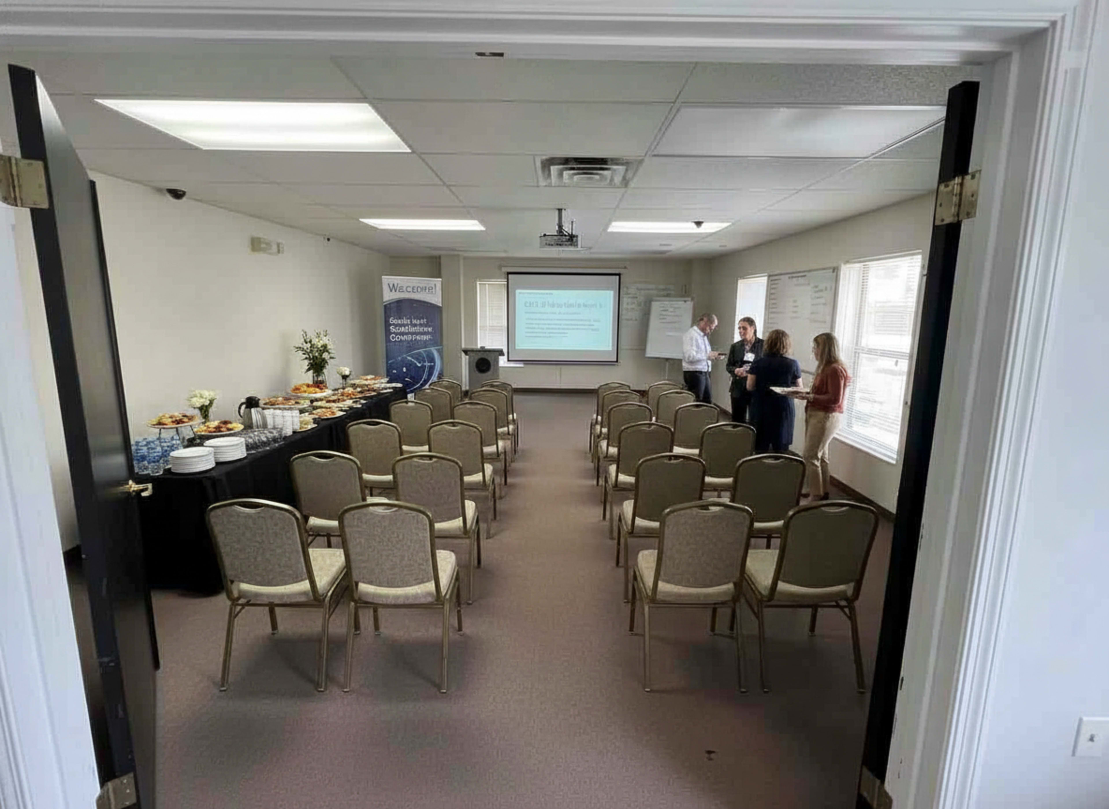 A meeting room is set up with rows of chairs facing a presentation screen, and a table with refreshments is positioned along one wall.