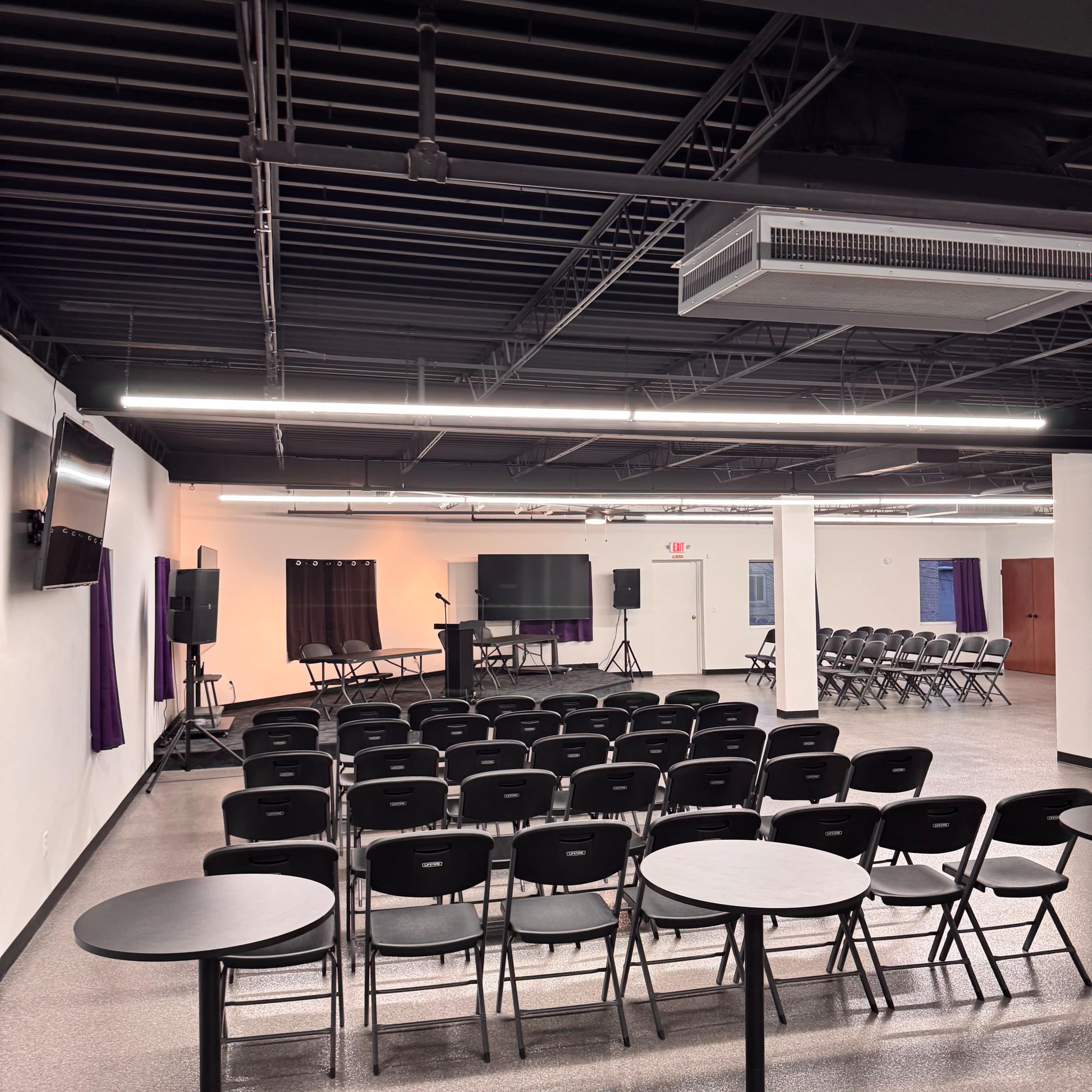 The image shows a large, empty meeting room with rows of black chairs facing a stage area, complete with audio-visual equipment and tables positioned around the space.