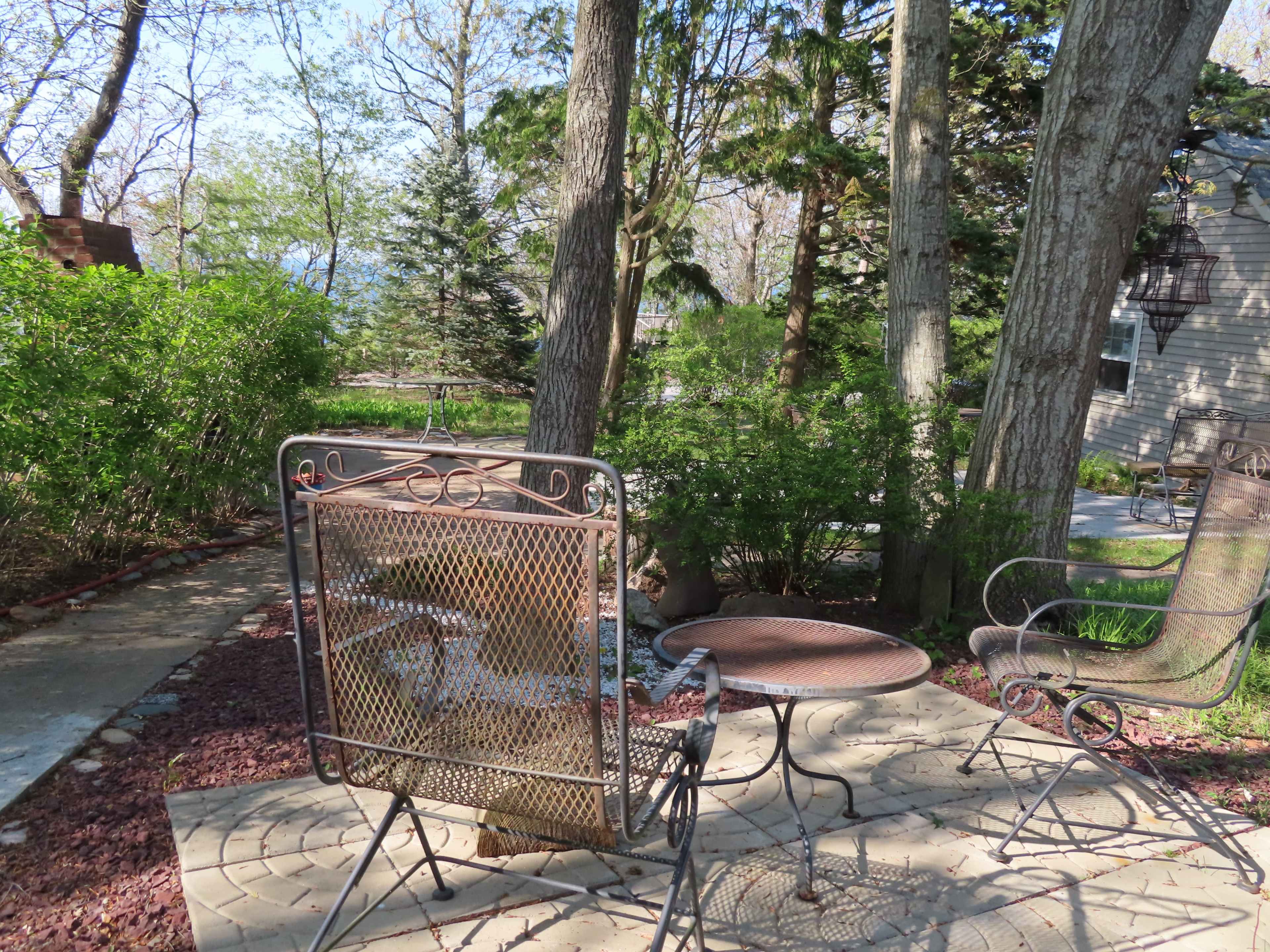A patio area features a metal chair and a small table surrounded by trees and shrubs.