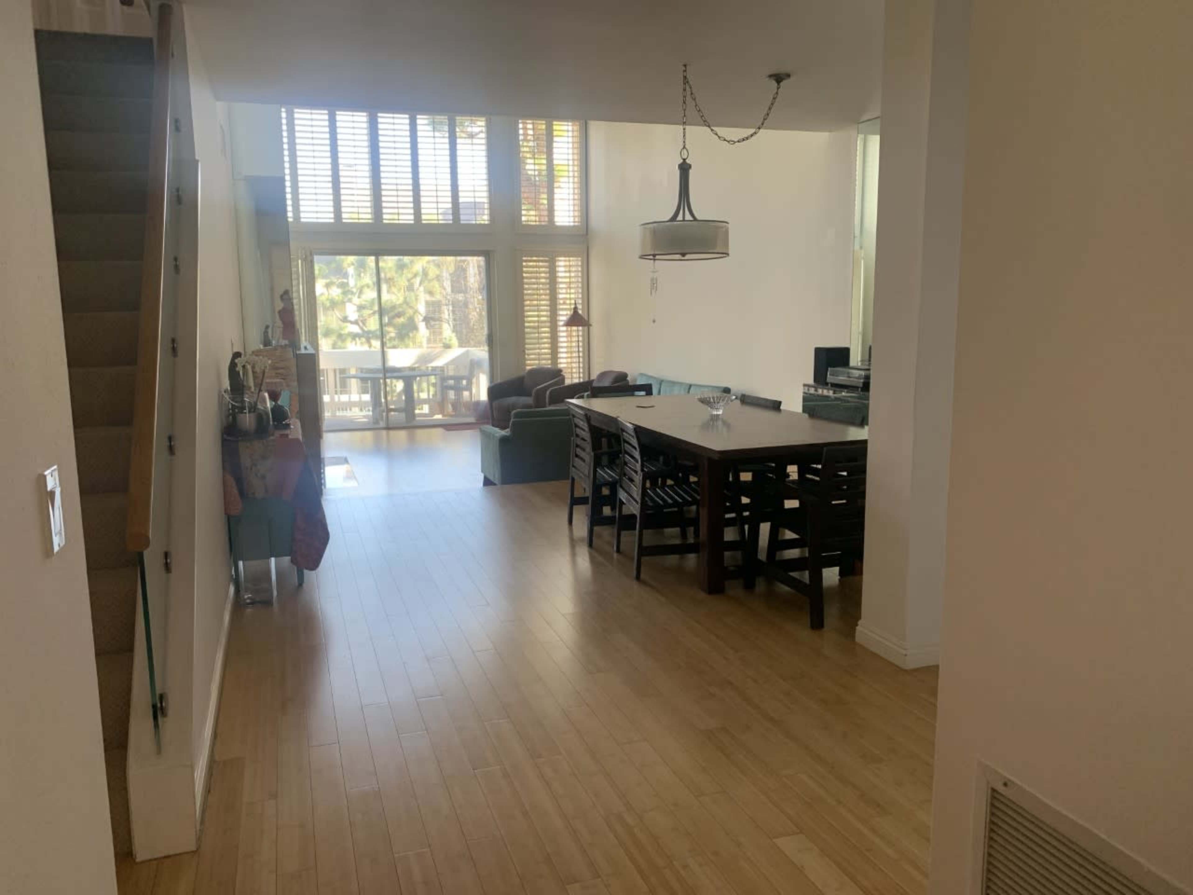 A spacious interior with a staircase on the left, a dining table in the center, and a living area with large windows in the background.
