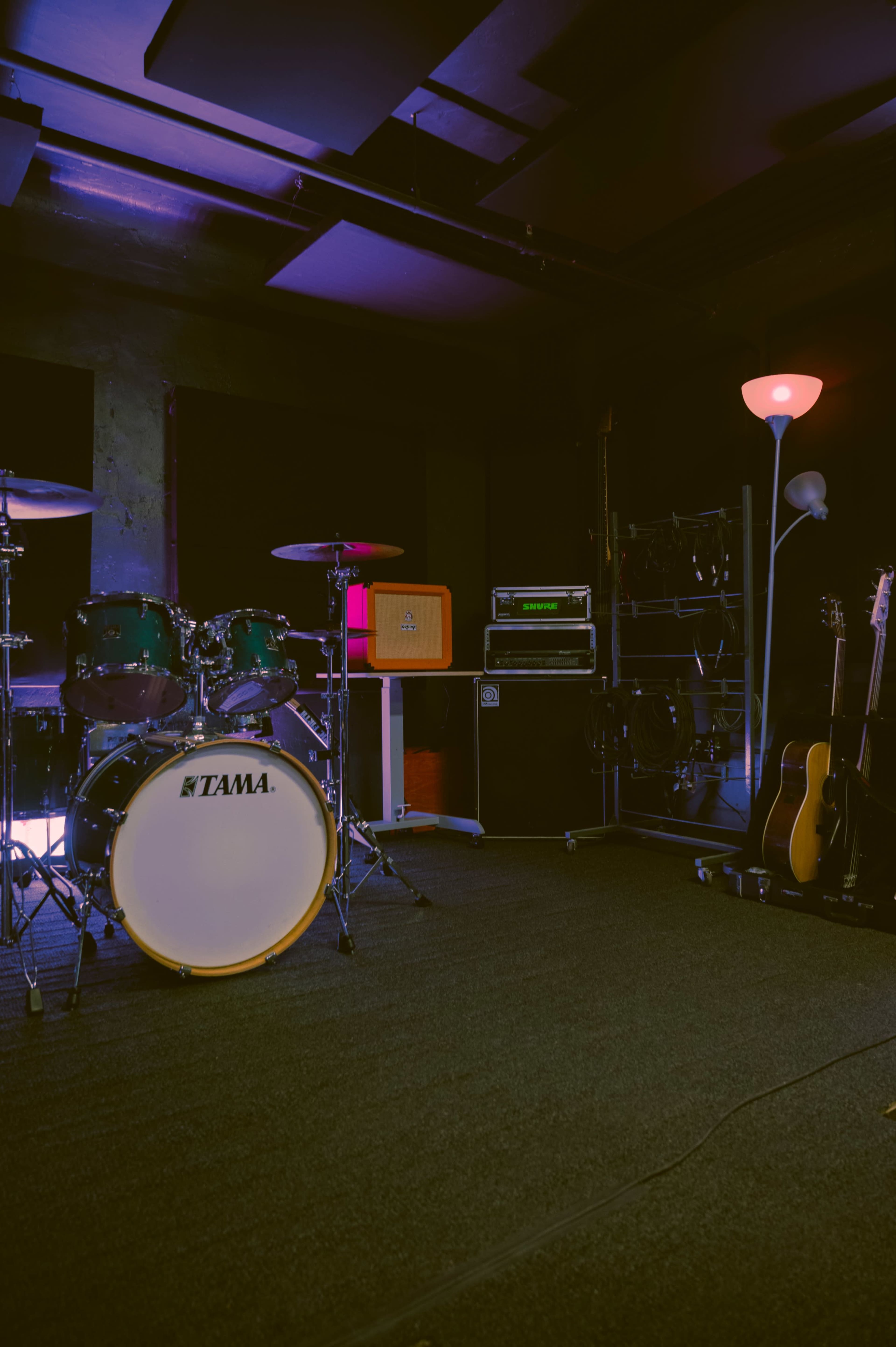 The image shows a music rehearsal space featuring a drum set, amplifiers, and a floor lamp against a dark wall.