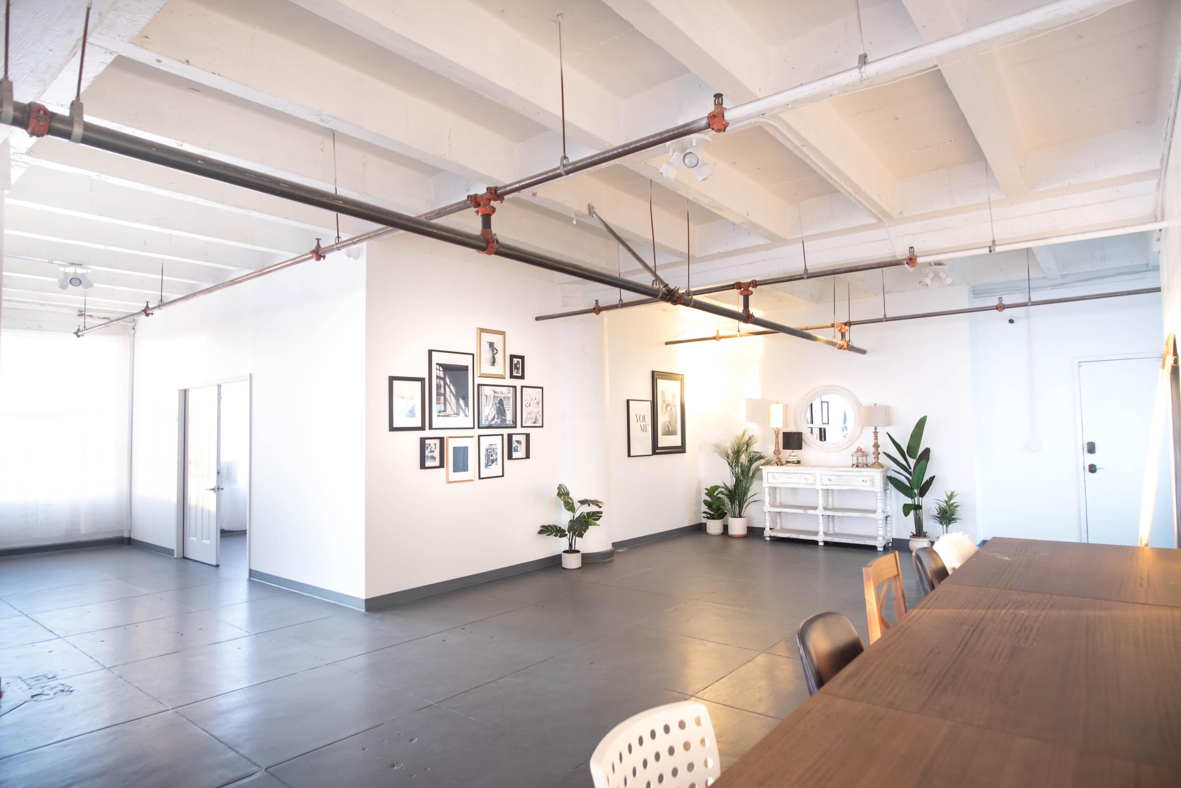 A spacious, minimalist interior with a long wooden table, potted plants, wall art, and large windows allowing natural light.