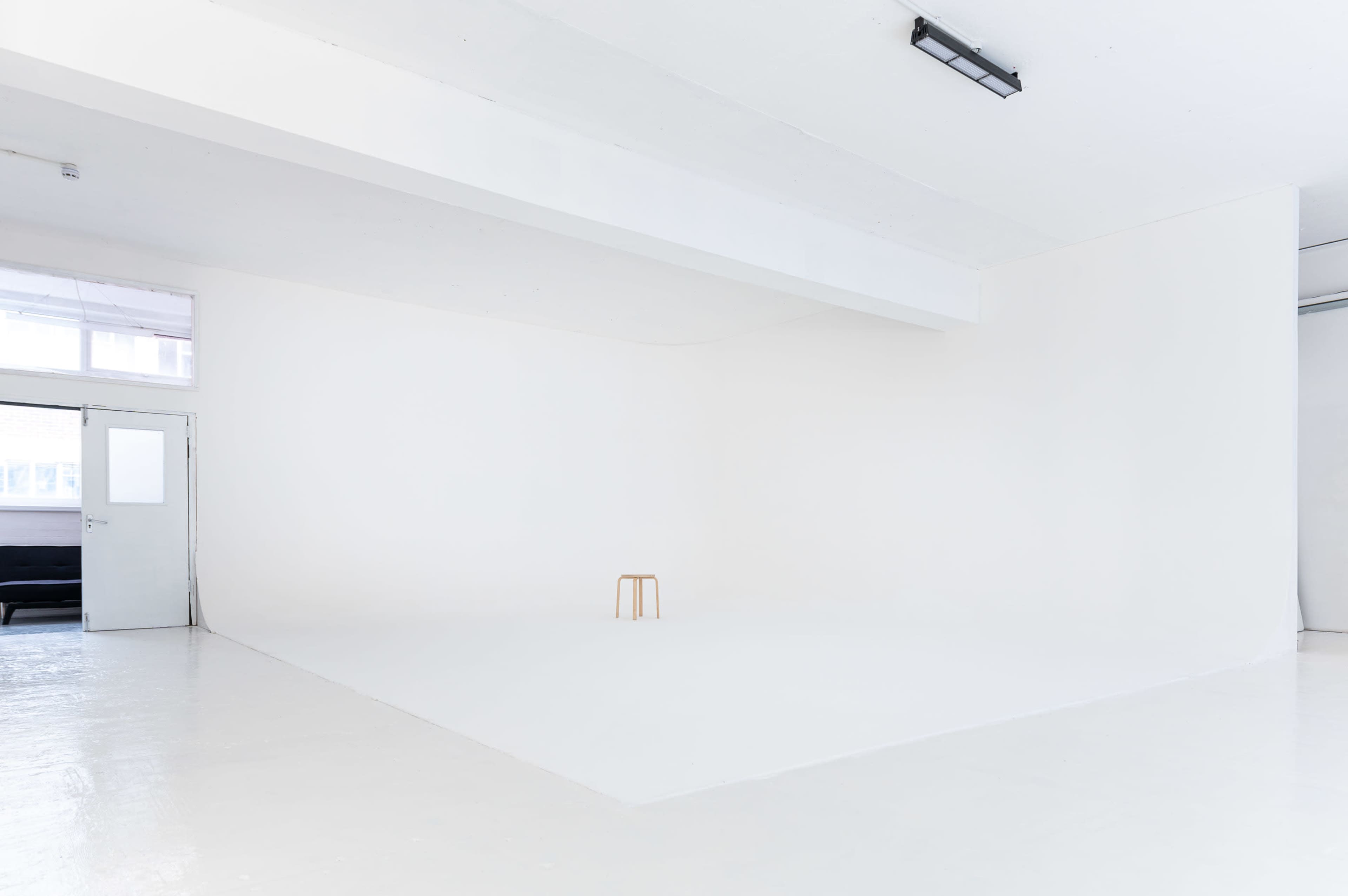 A sparsely furnished white room features a single wooden stool in the center.