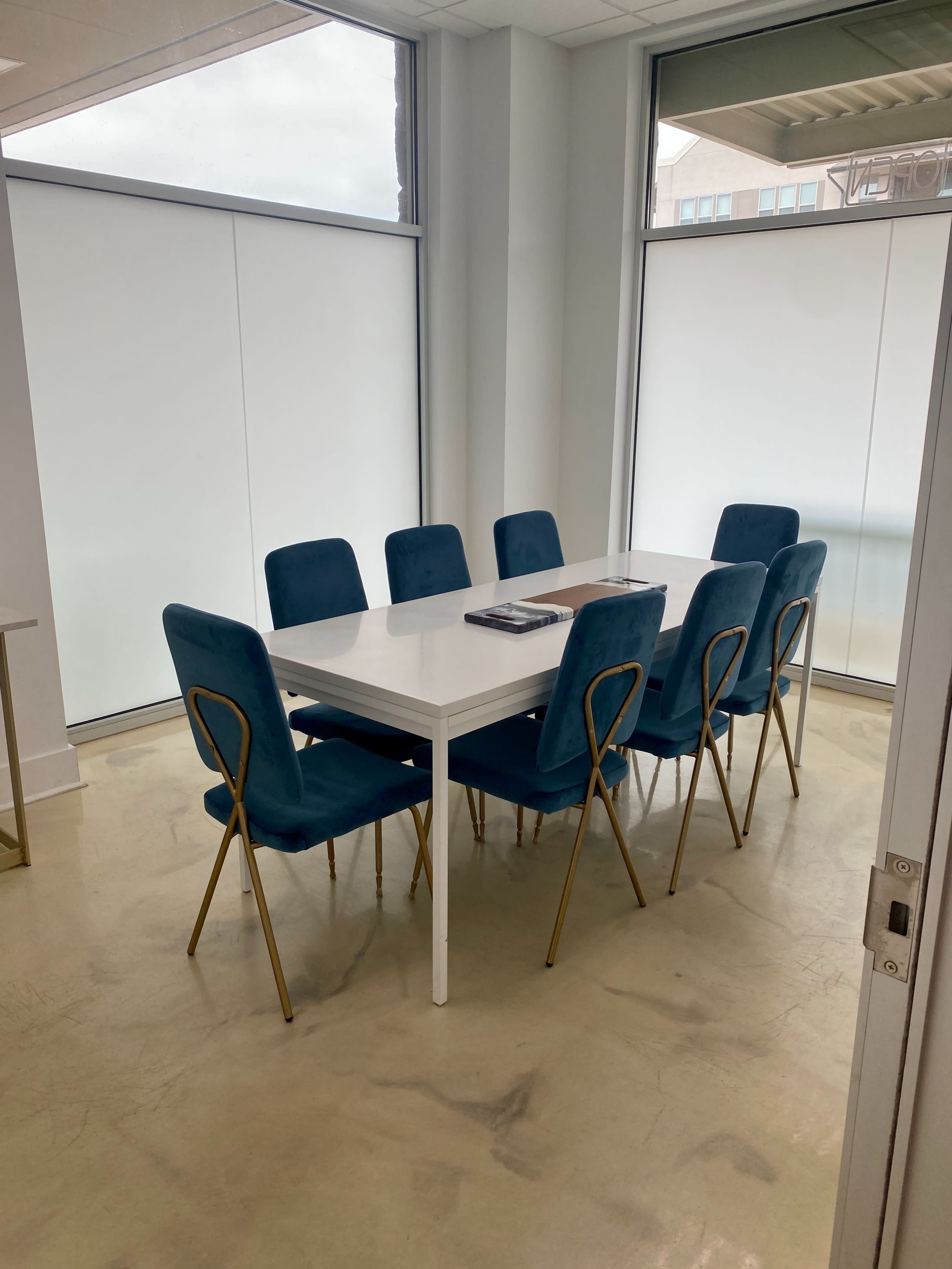 A bright meeting room features a white rectangular table surrounded by eight blue chairs with gold legs, situated near large windows.