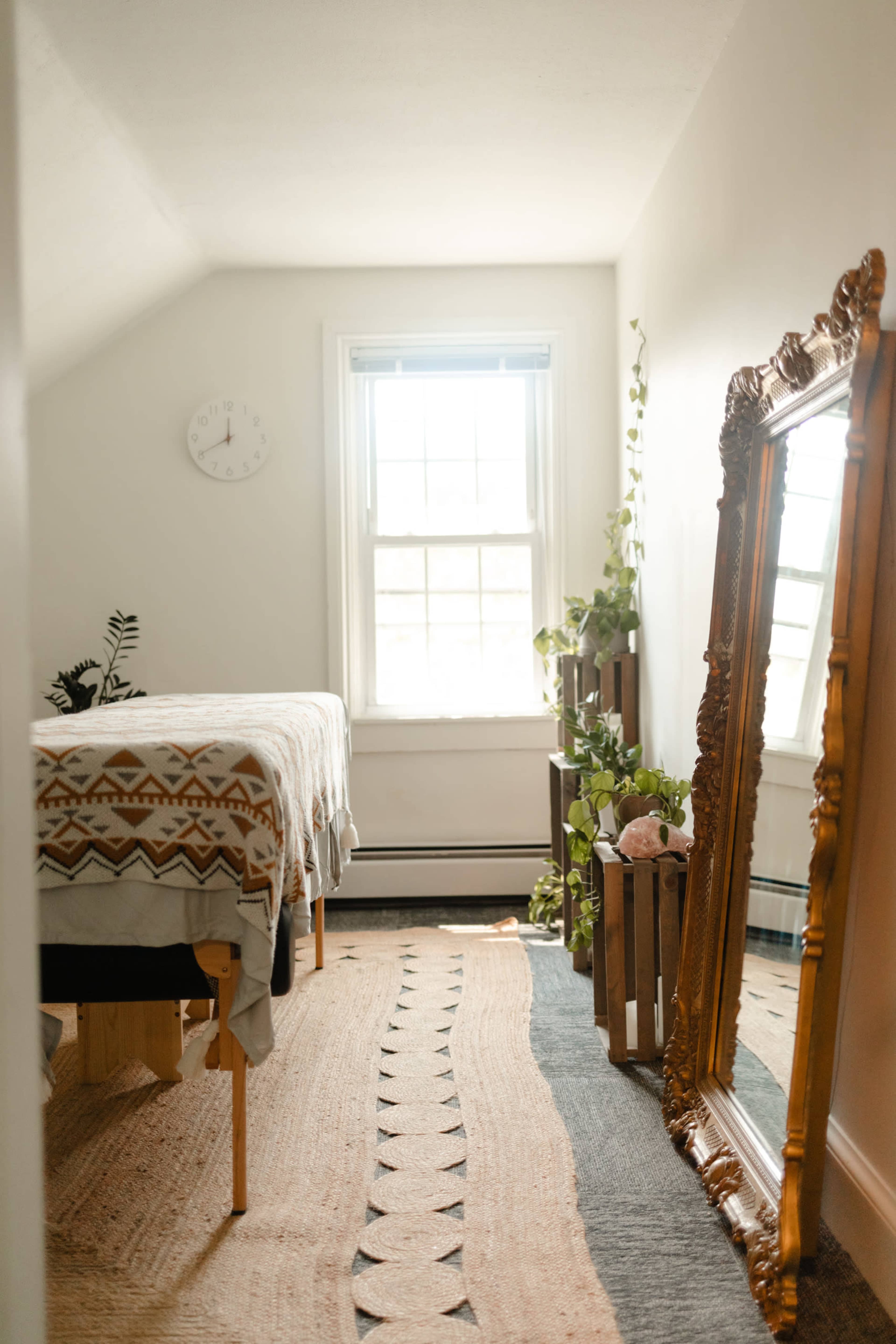 A bright bedroom with a bed covered by a patterned blanket, a large mirror, and plants arranged around a window.