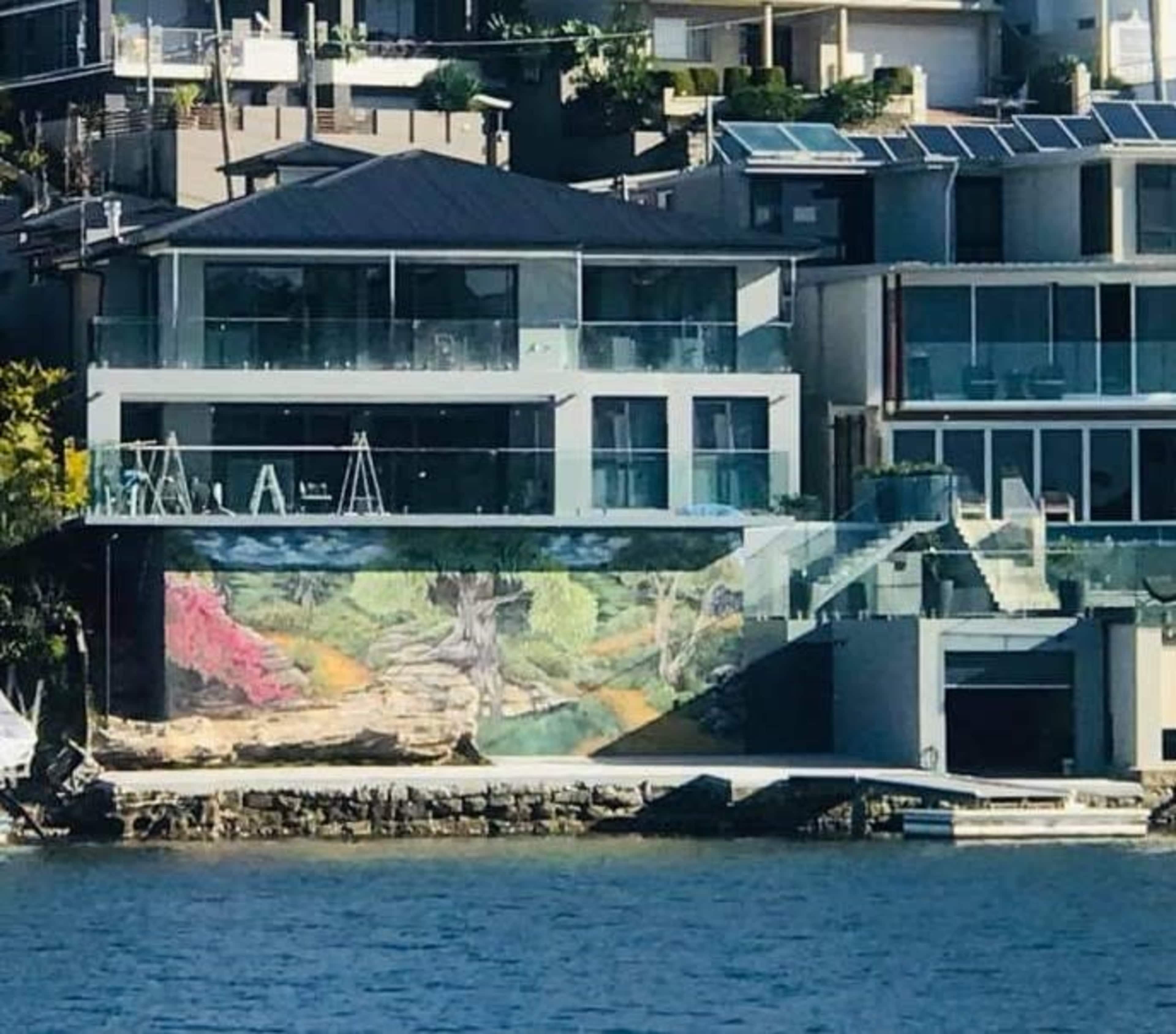 The image shows a waterfront property featuring a large mural on its wall, surrounded by modern houses and greenery.