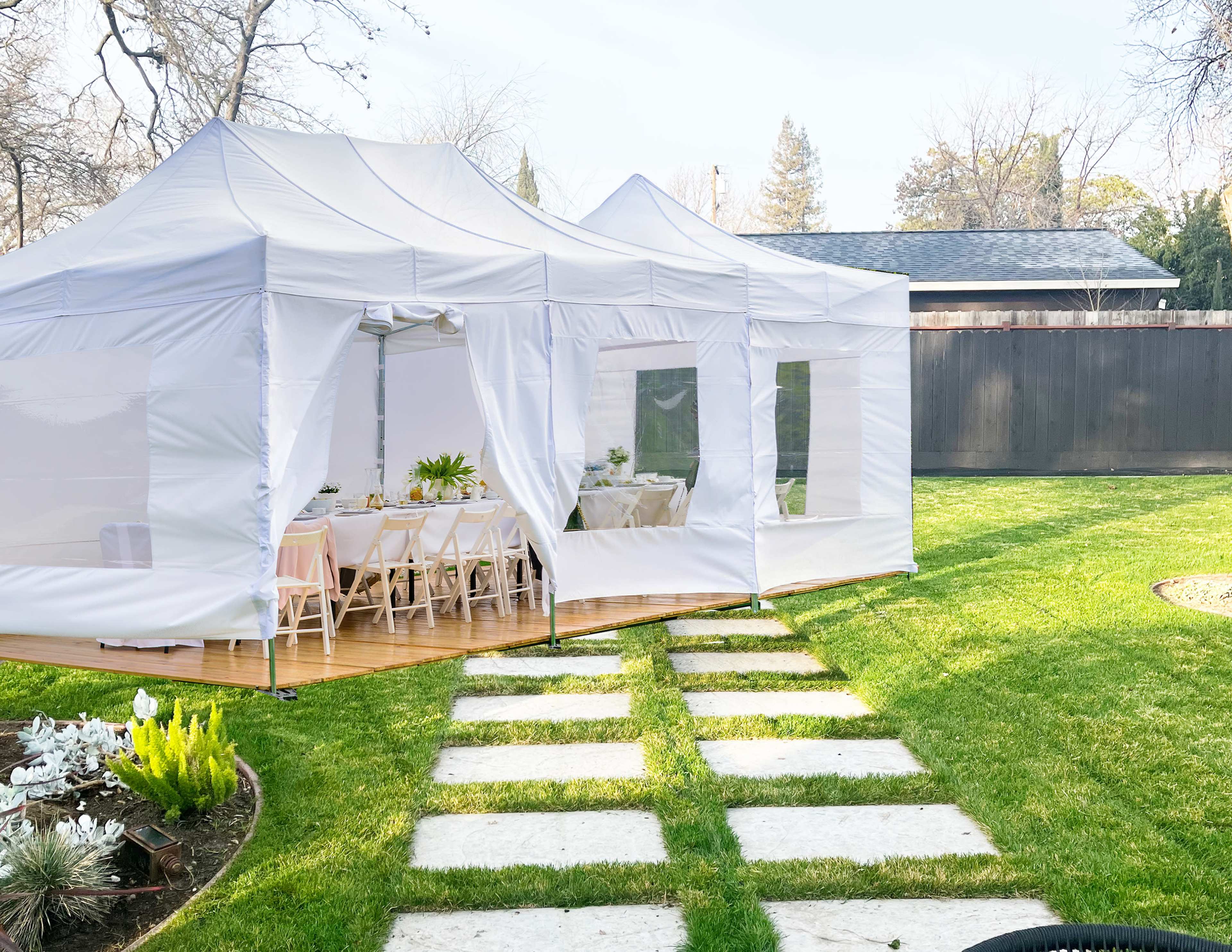 A white outdoor tent with several tables is set up on a grassy area, surrounded by stone walkways.
