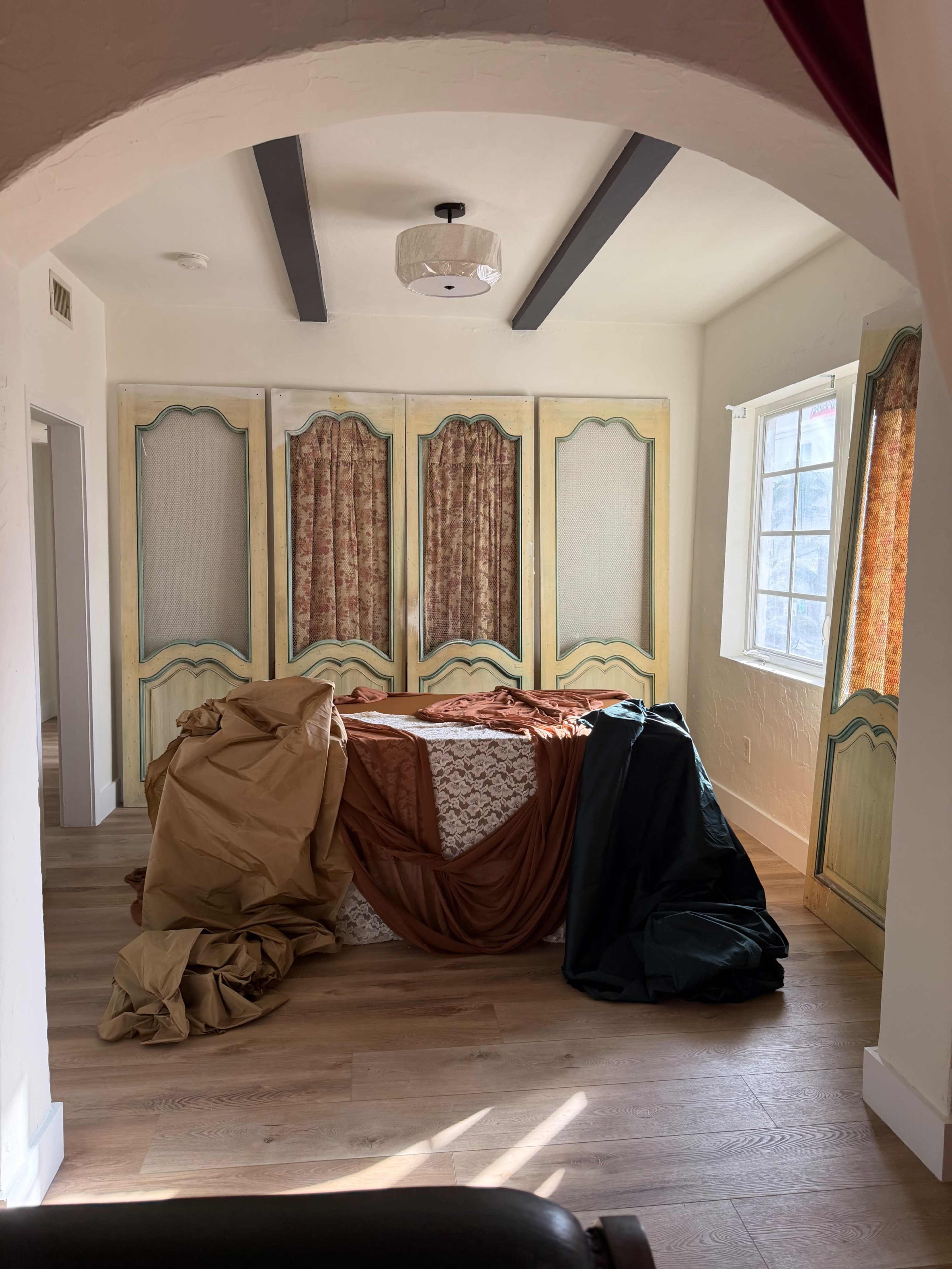 The image shows a room with a bed covered in fabric and draped materials, surrounded by patterned curtains and a decorative wooden screen.