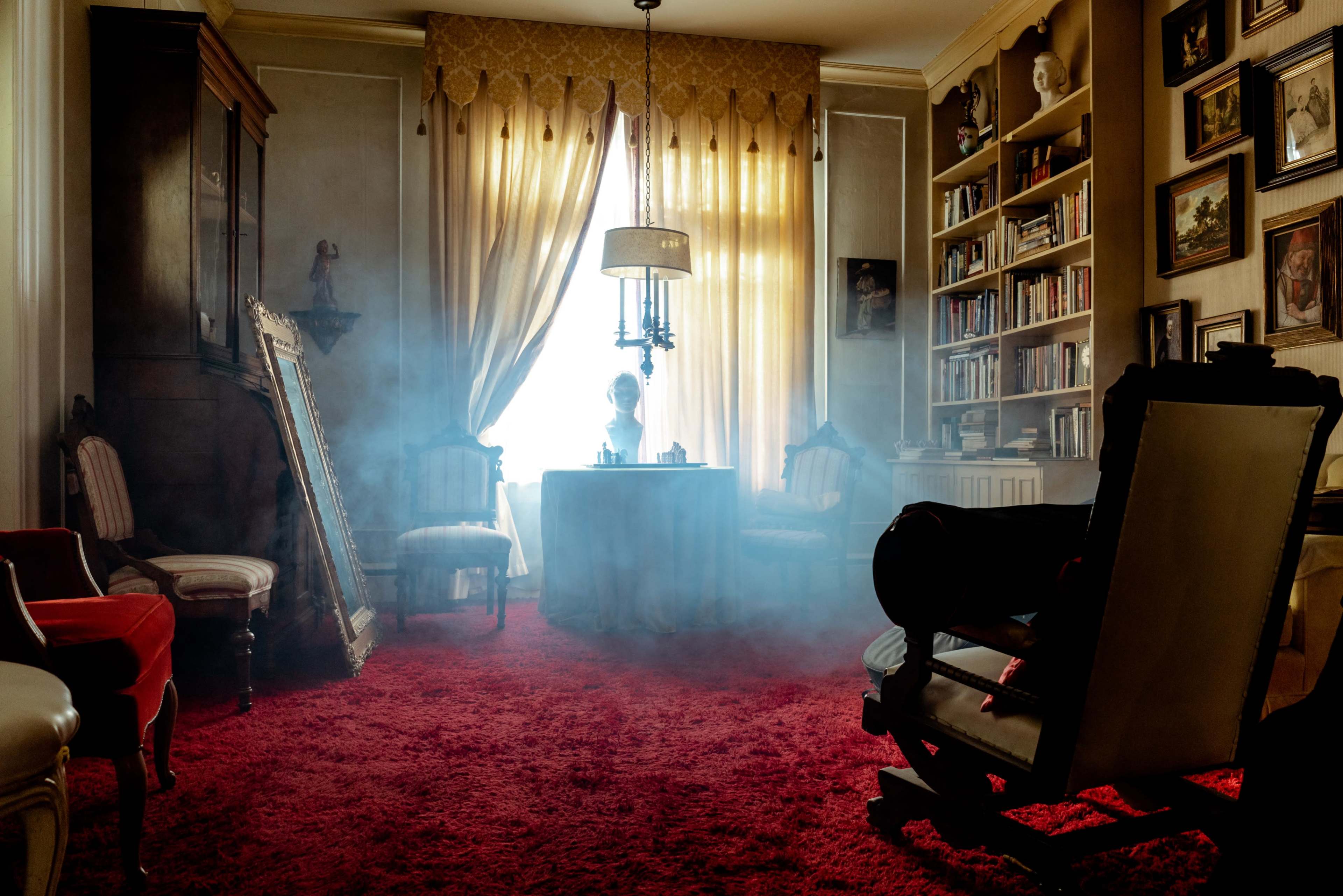 A dimly lit room features a round table surrounded by antique chairs, with smoke hovering in the air and a large window draped with golden curtains.
